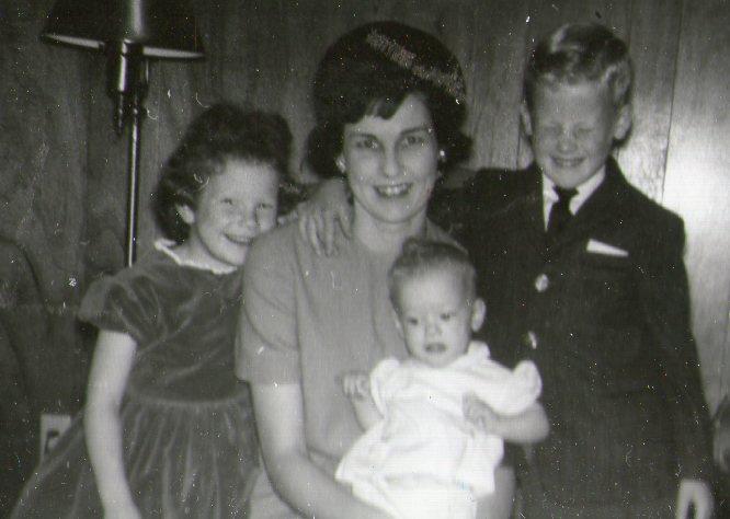 Group of four, smiling mother with three children in retro clothing posing indoors by a lamp.