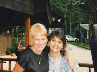 Two friends enjoy each other's company outside, smiling warmly amidst green trees and sunlight.