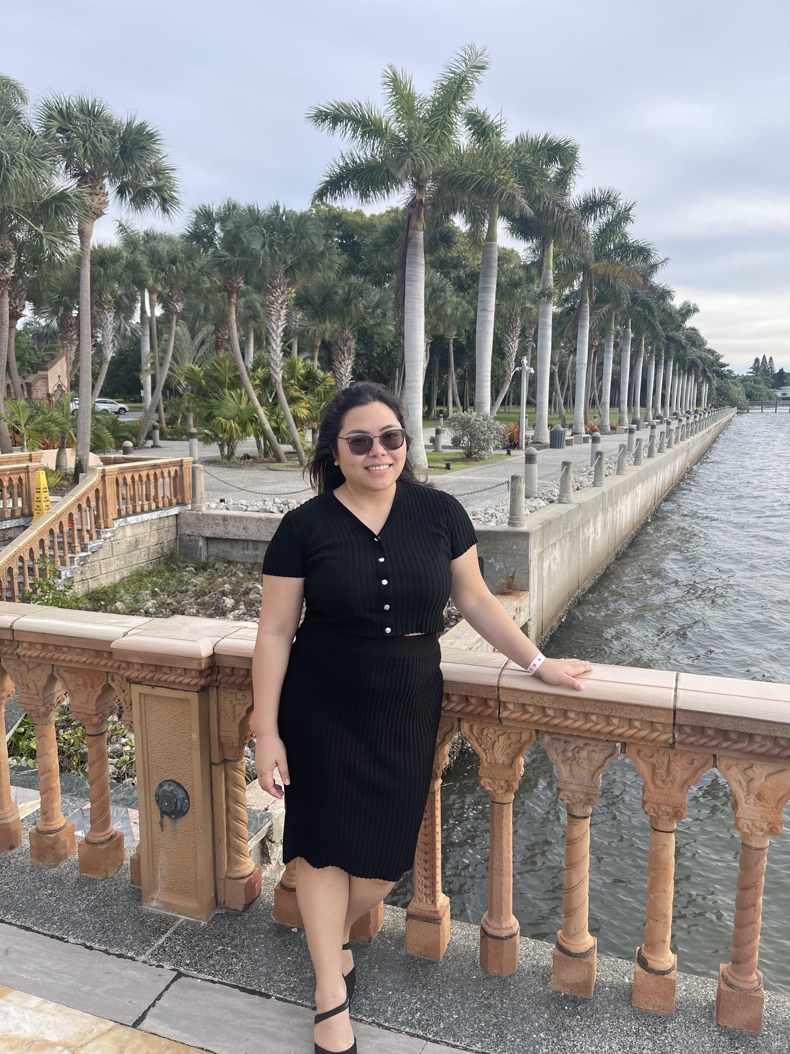 A woman stands on a decorative railing by the water, surrounded by palm trees and greenery.
