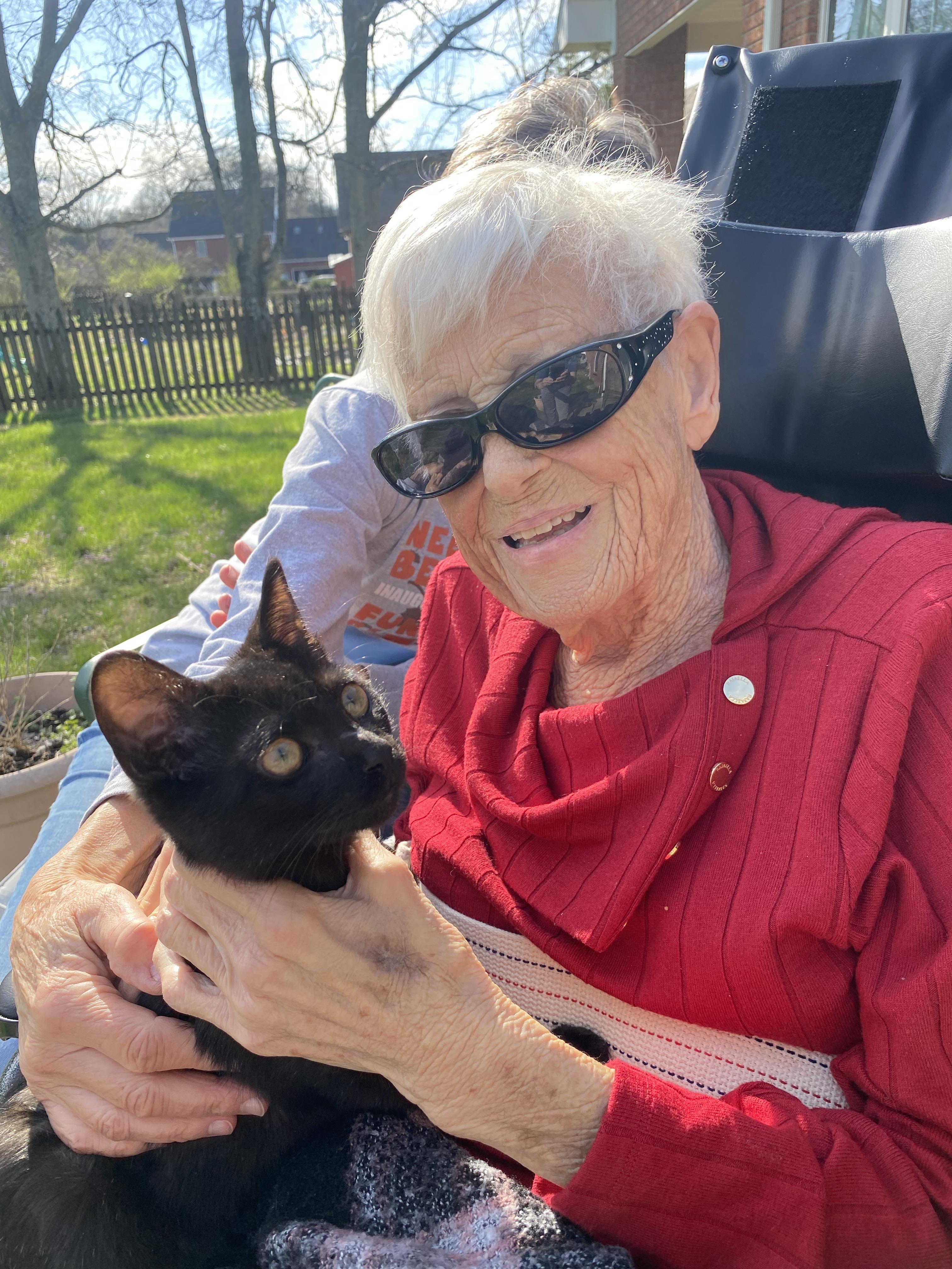 Smiling elderly woman sits outside, holding a black cat in her lap while enjoying the warm sun.
