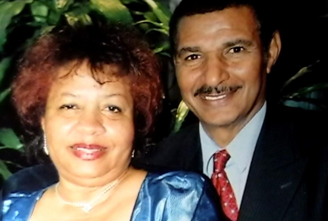A joyful couple dressed elegantly smiles for the camera in a lively atmosphere filled with plants.