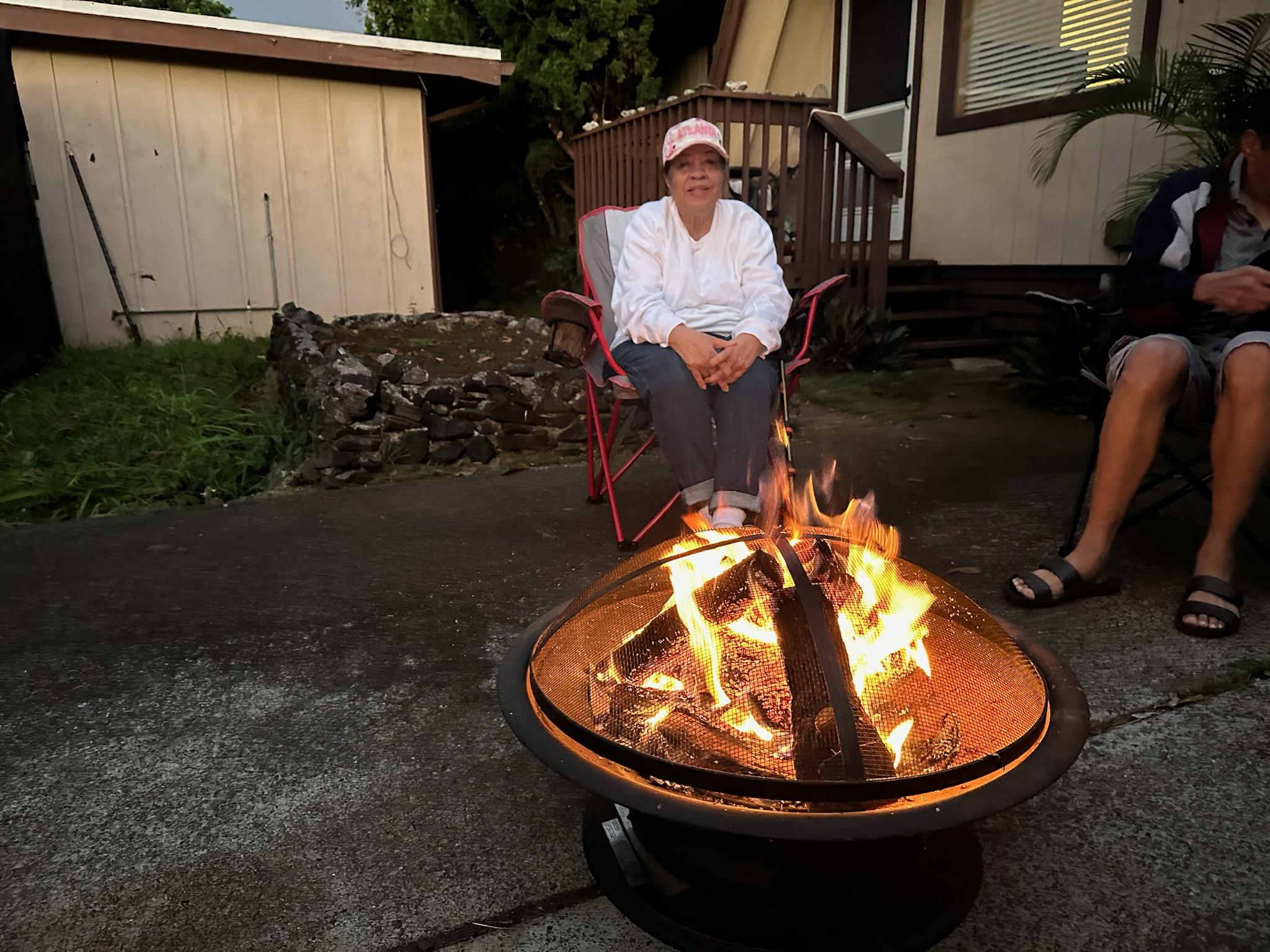 A woman sits comfortably by a fire pit wearing casual attire, enjoying a serene night with family.