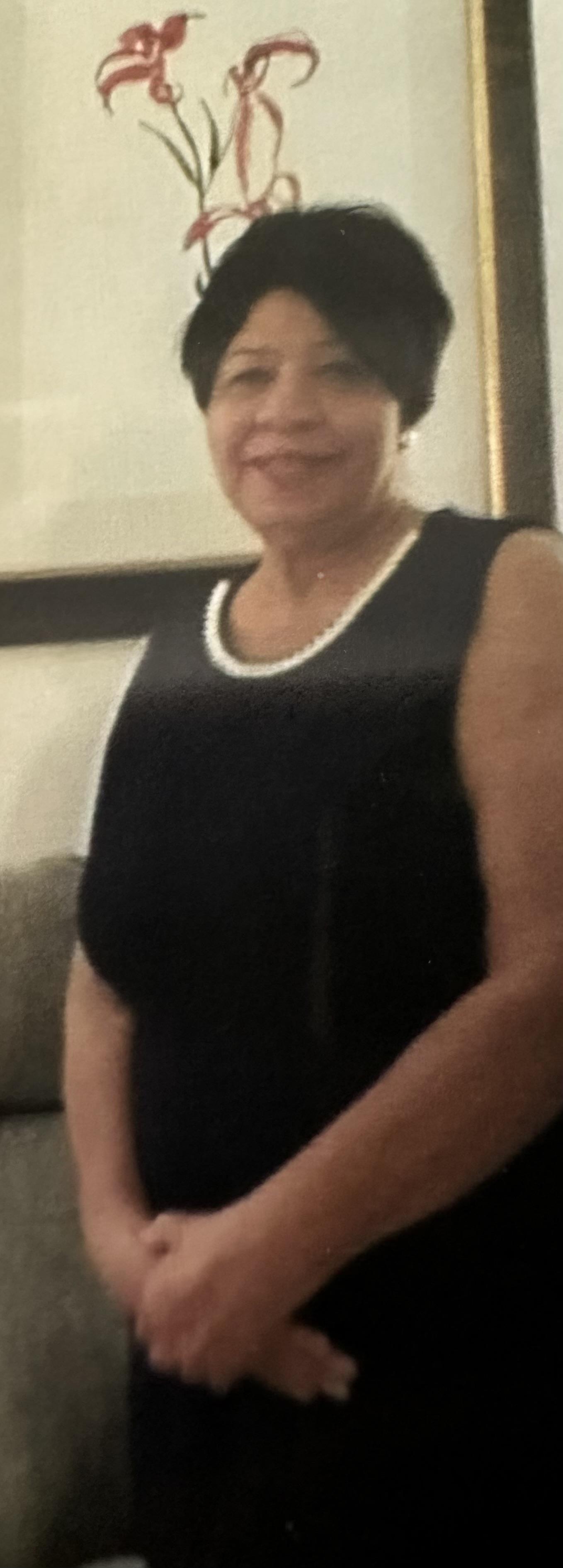 A woman dressed in a black outfit with a pearl necklace poses beside a sofa in a cozy room.