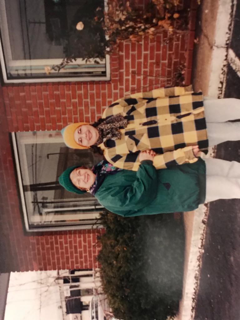 Friends stand outside a brick house wearing bright winter attire, smiling in the cold weather.