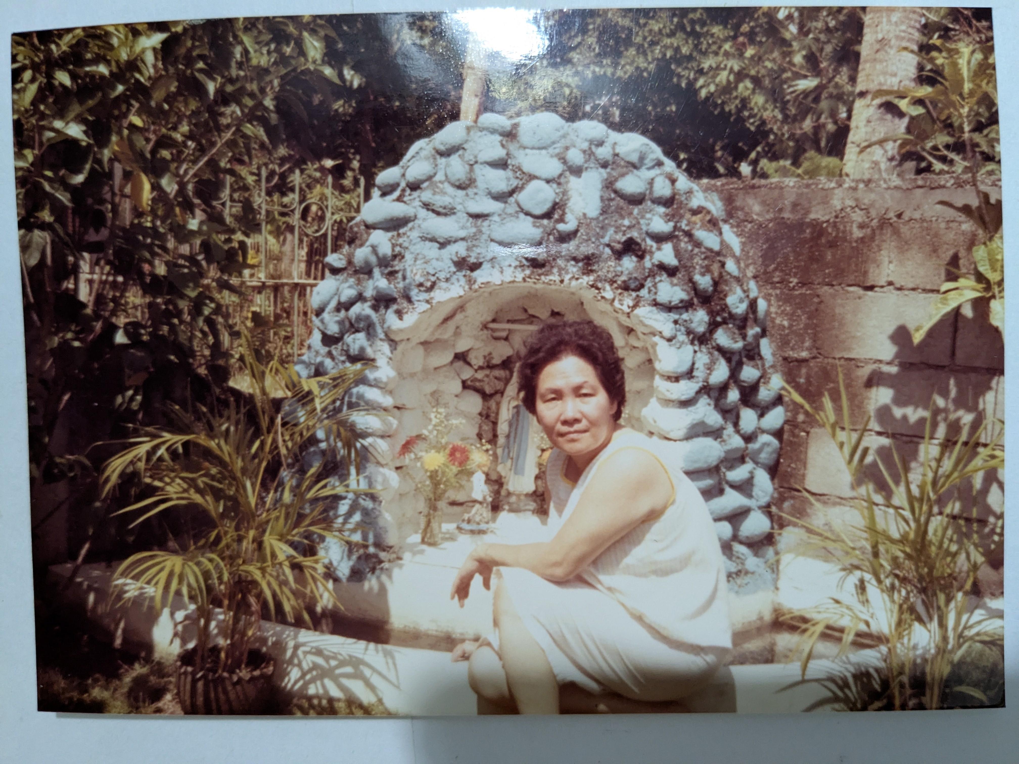 A woman poses gracefully next to a unique stone structure, surrounded by greenery.