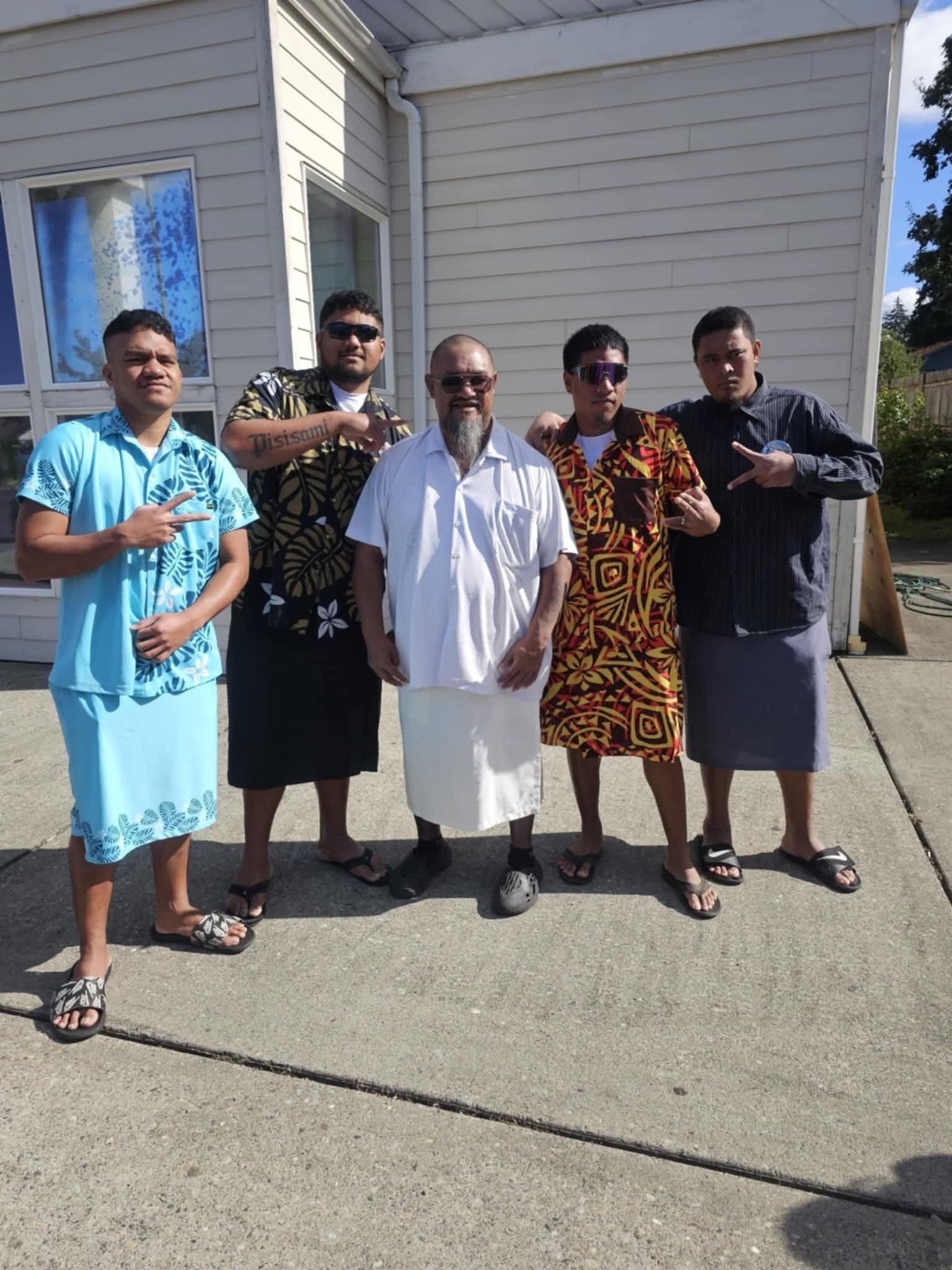 Five men are gathered in front of a house, smiling and wearing traditional clothing.