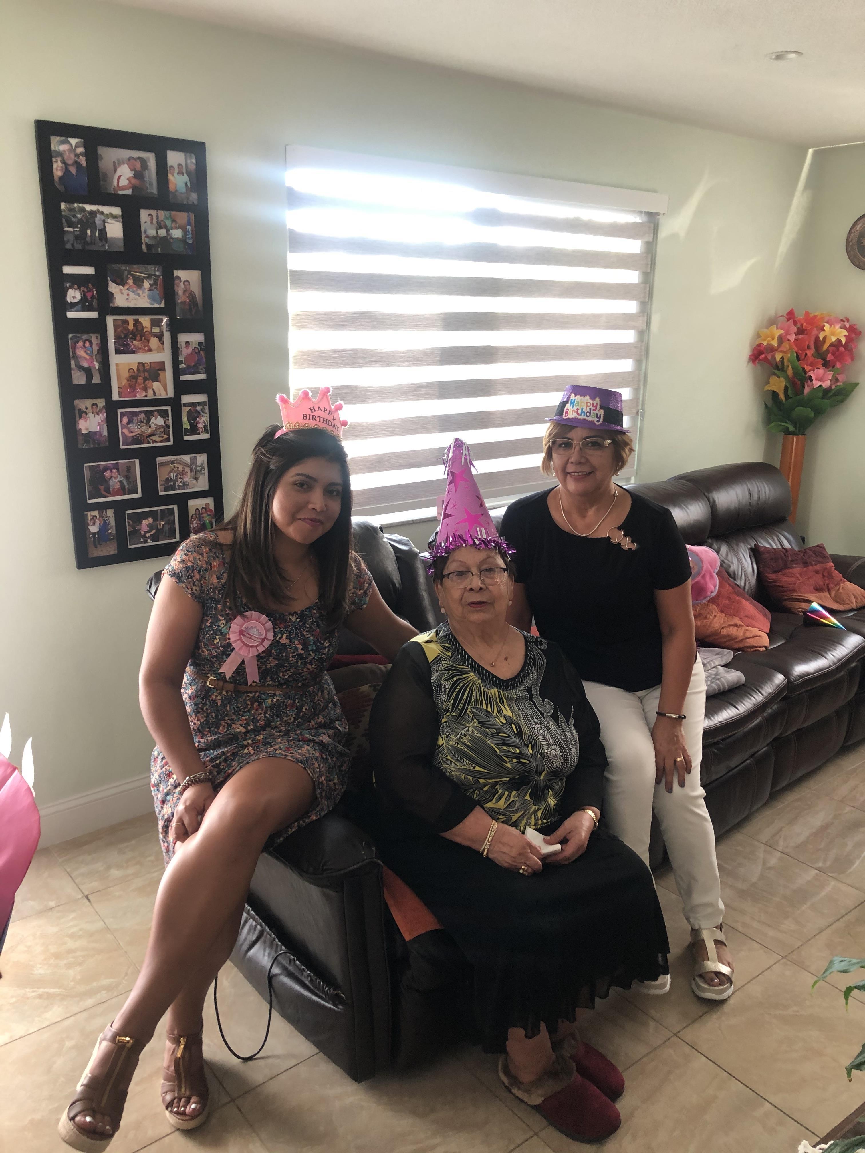 Women gather in a cheerful living room for a birthday celebration, wearing festive hats and smiling.