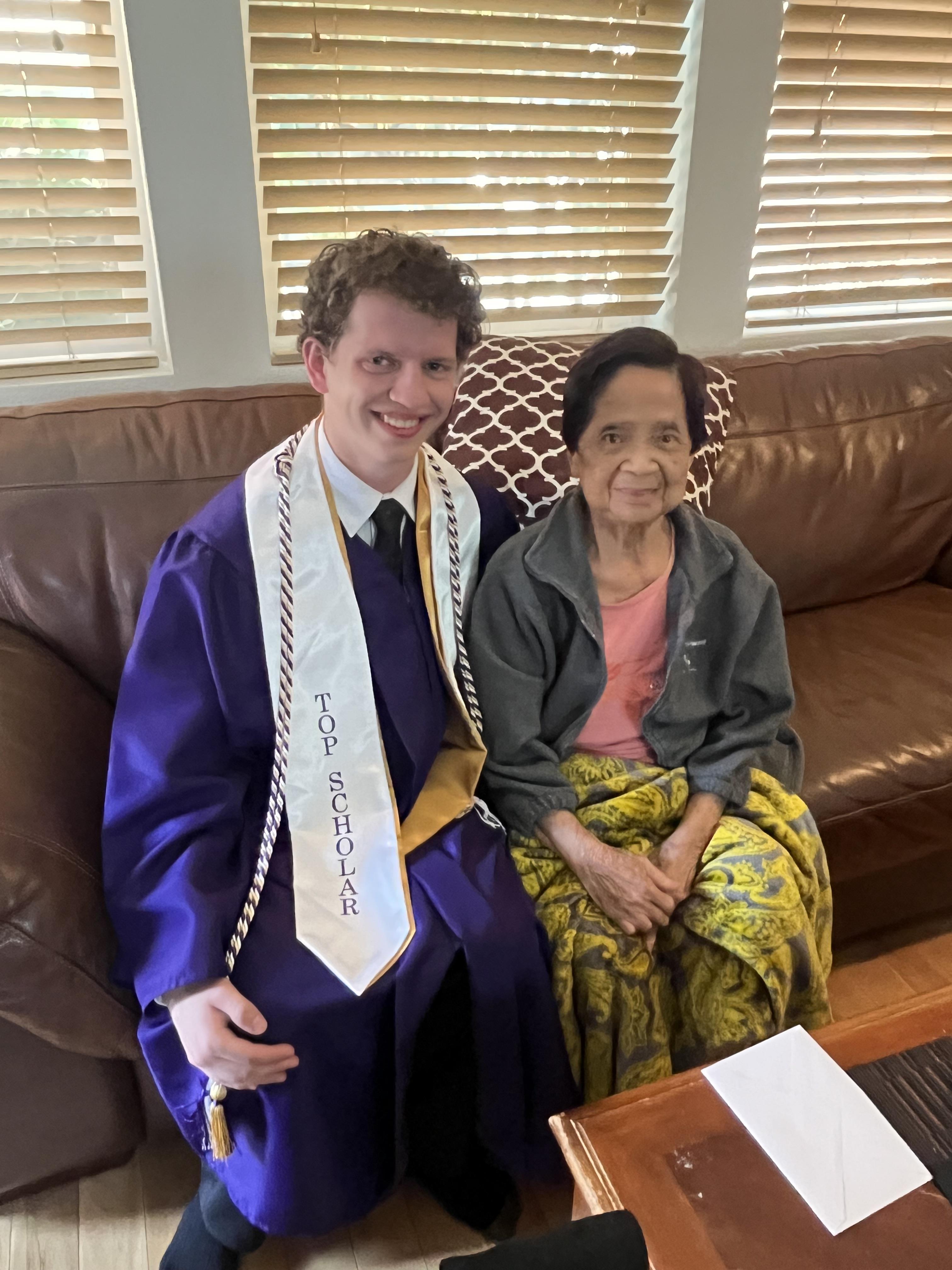 A graduate in a cap and gown proudly poses with an older woman on a couch, celebrating their bond.