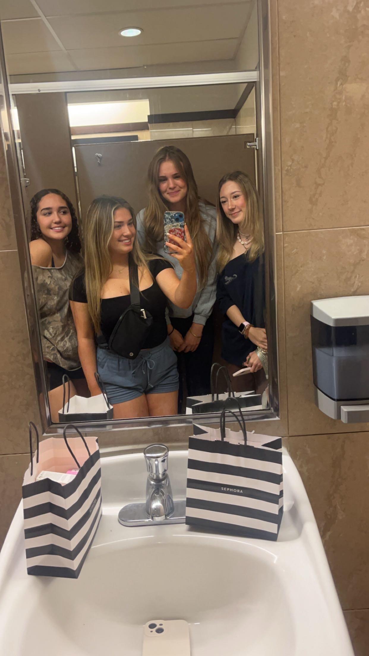 Four friends pose together for a selfie in a well-lit bathroom while shopping.