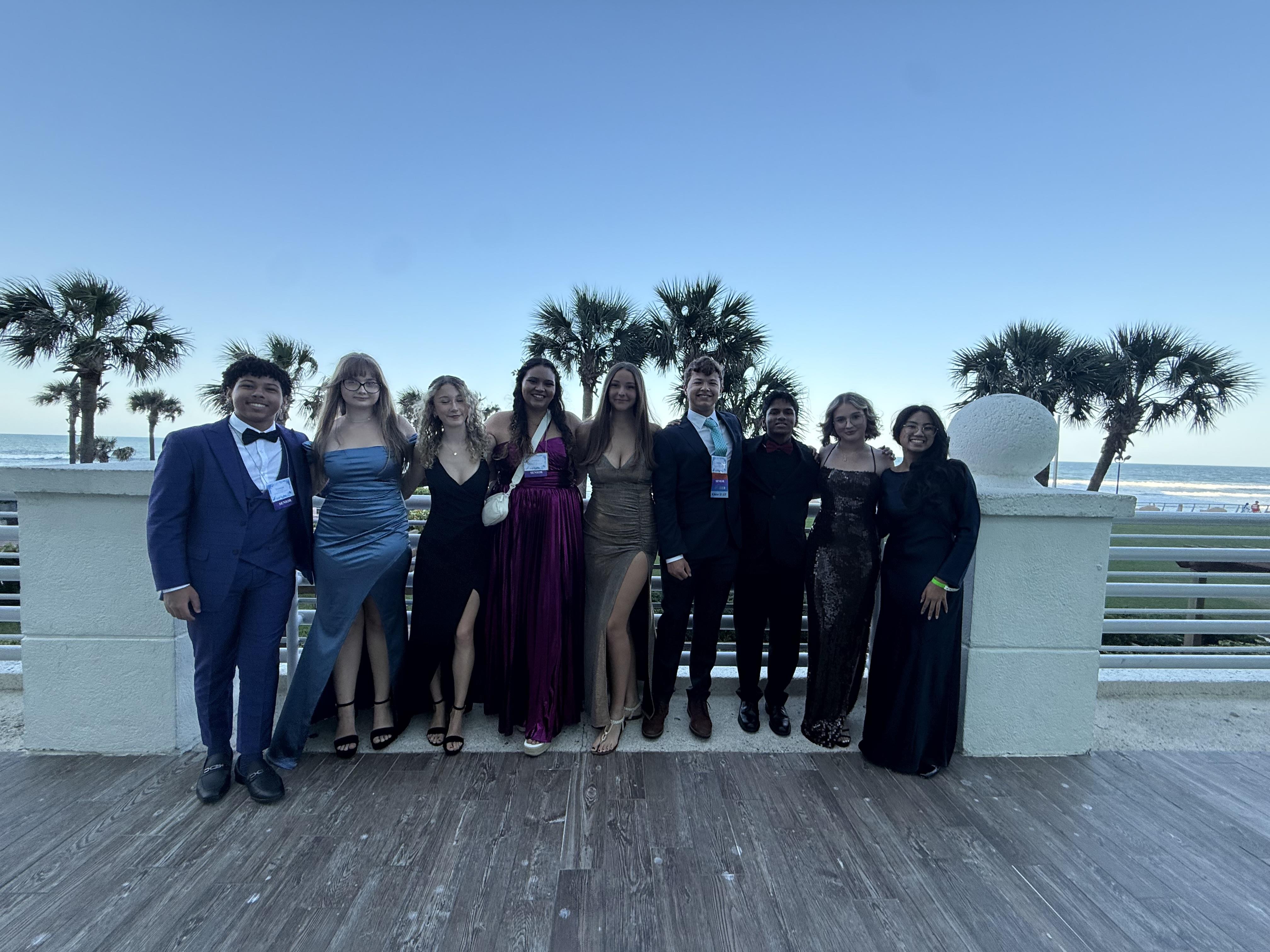 Formally dressed group enjoys a moment together on a coastal deck as the sun sets in the background.