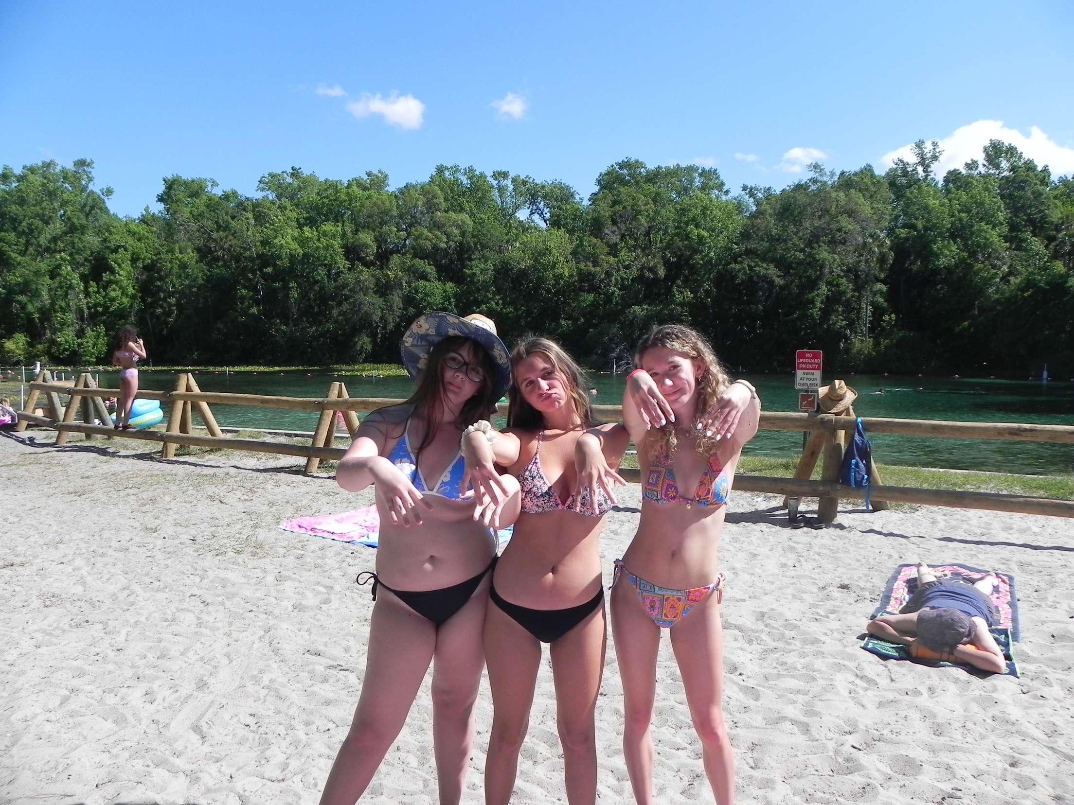 Three girls wearing swimwear playfully pose on the sandy beach under a clear sky.