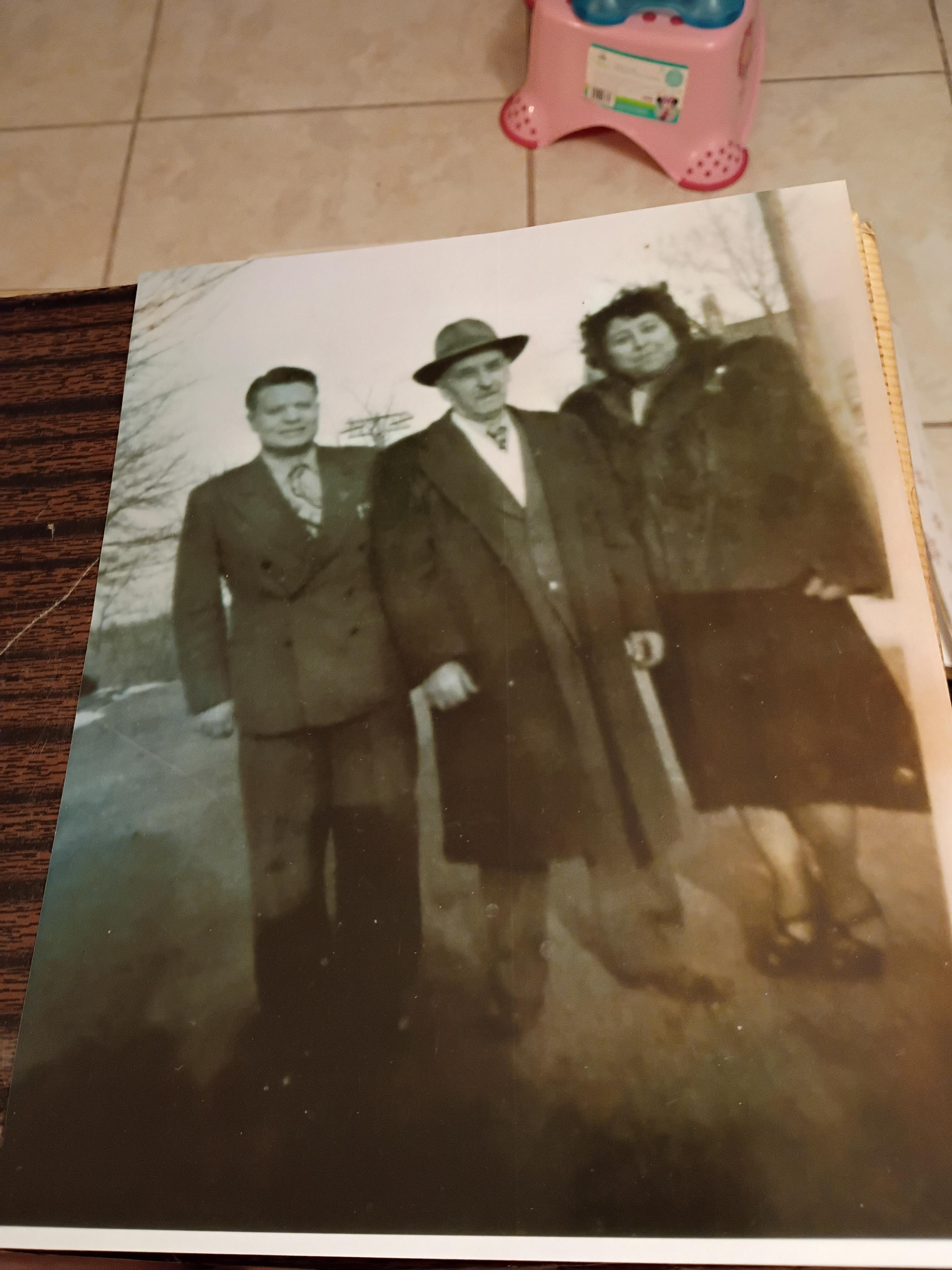 Three individuals in vintage formal wear pose outdoors in the sunlight, amidst nature.