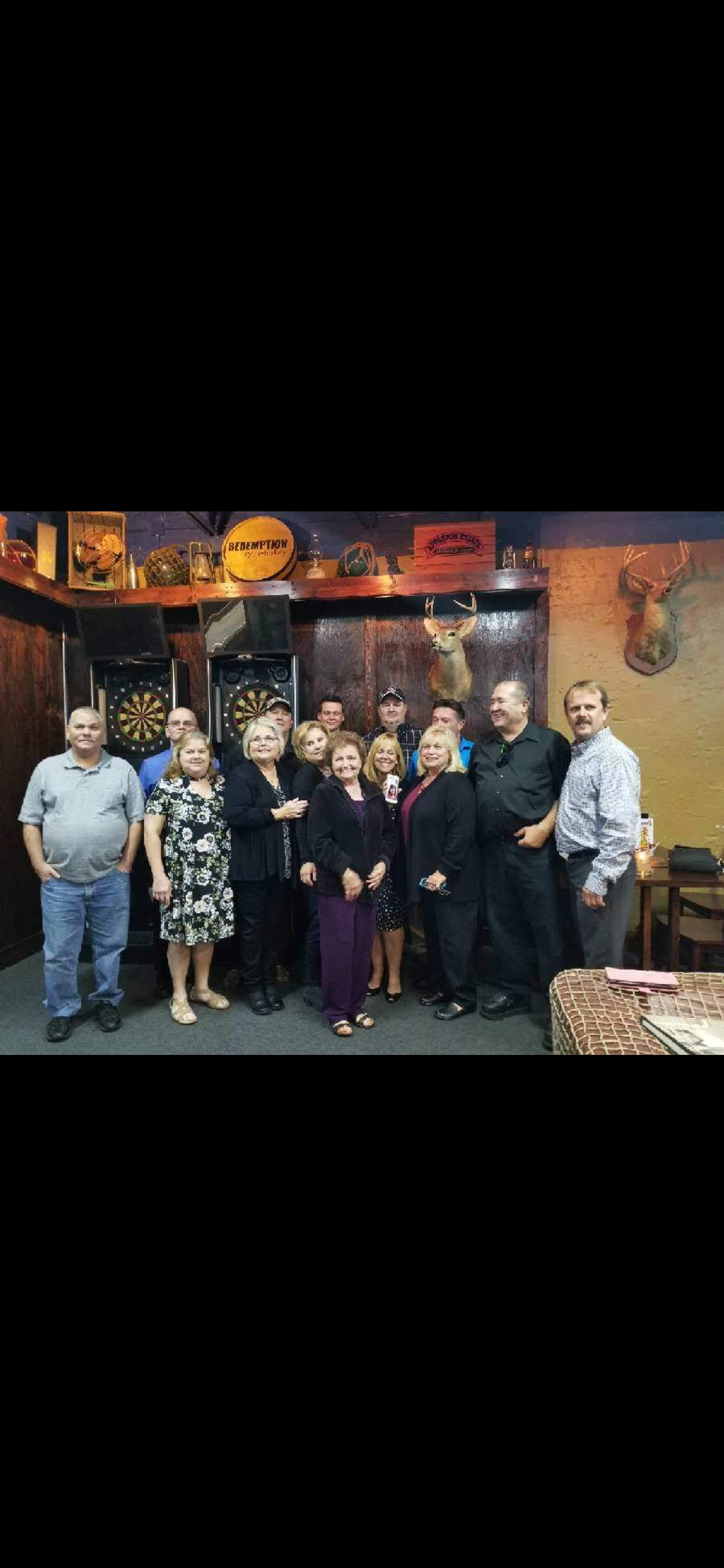 A group of friends poses together, smiling, inside a rustic bar with dartboards and wood decor.