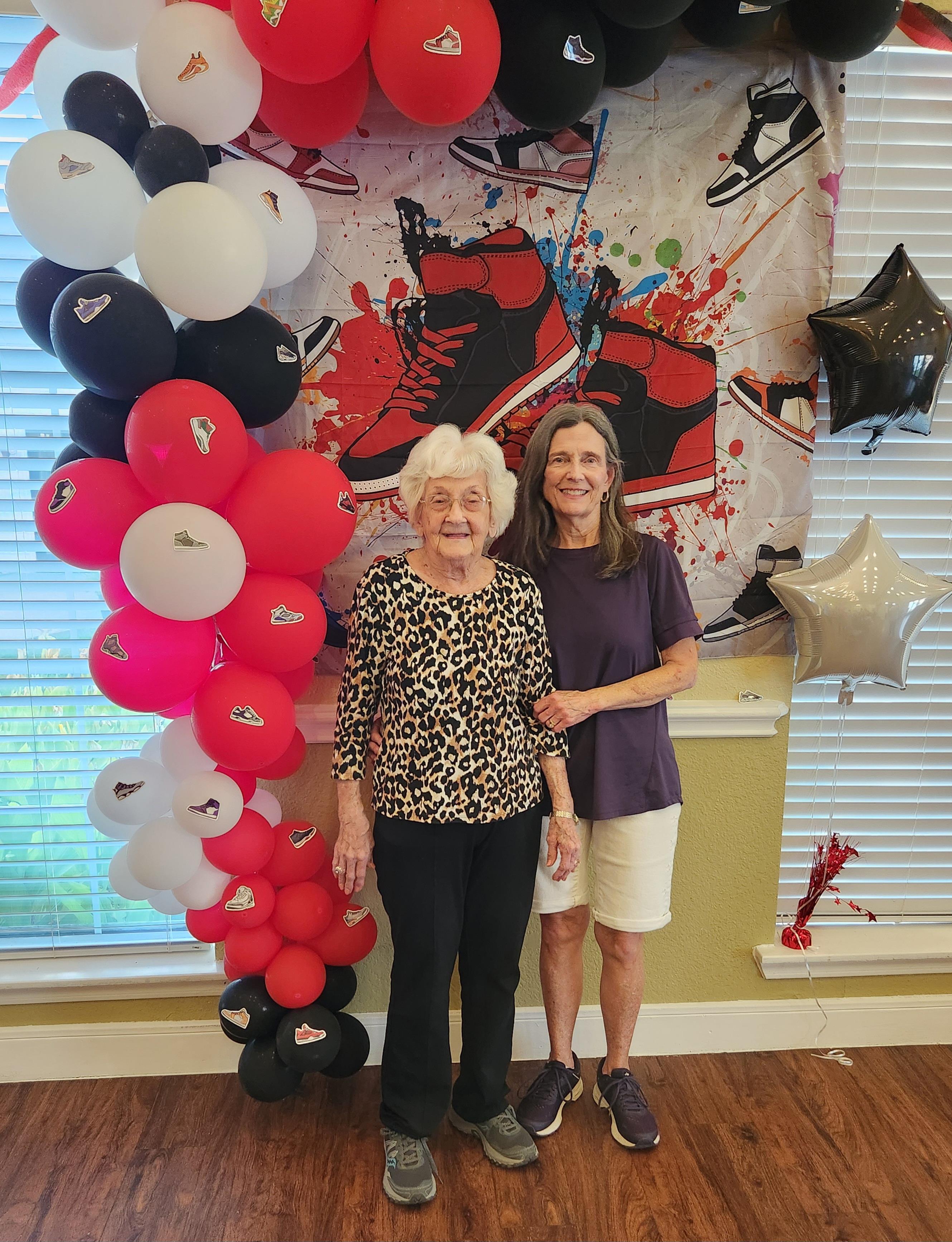 Two women stand together, smiling in front of a vibrant balloon arch at a joyful celebration.