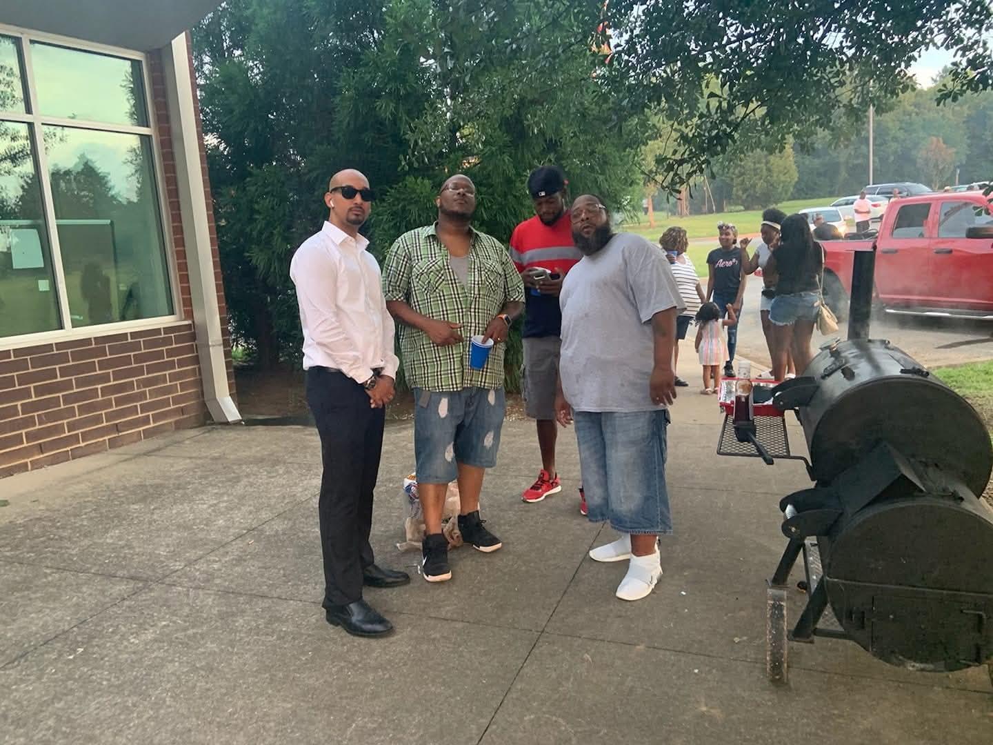 Four men engage in lively conversation on a warm day under the trees, enjoying their time together.
