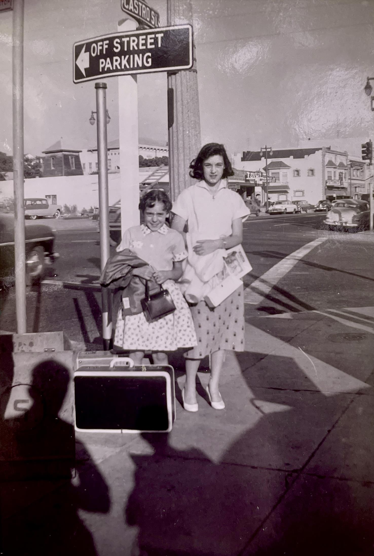 Two women stand at a city corner, one holding a suitcase, waiting for transport in the past.