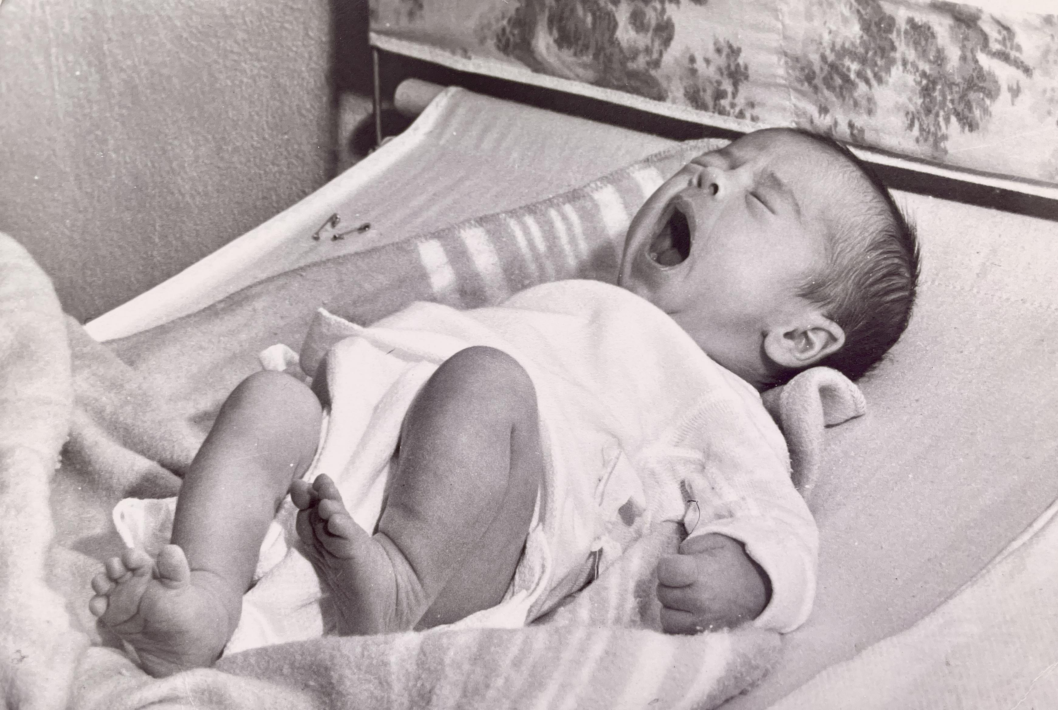 A baby in a cradle yawns widely while resting comfortably, bathed in soft afternoon light.