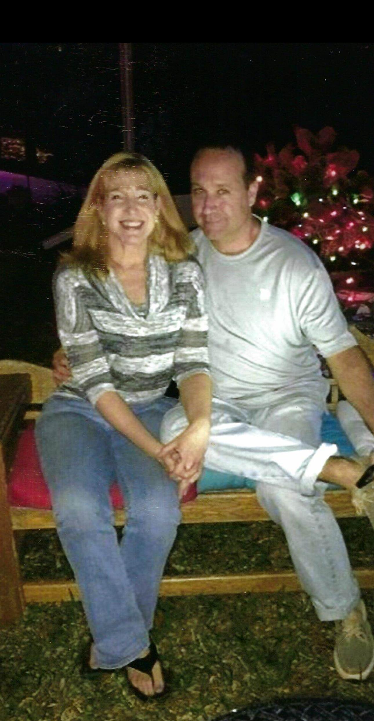 A happy couple sits closely on a wooden bench surrounded by colorful lights, sharing laughter.