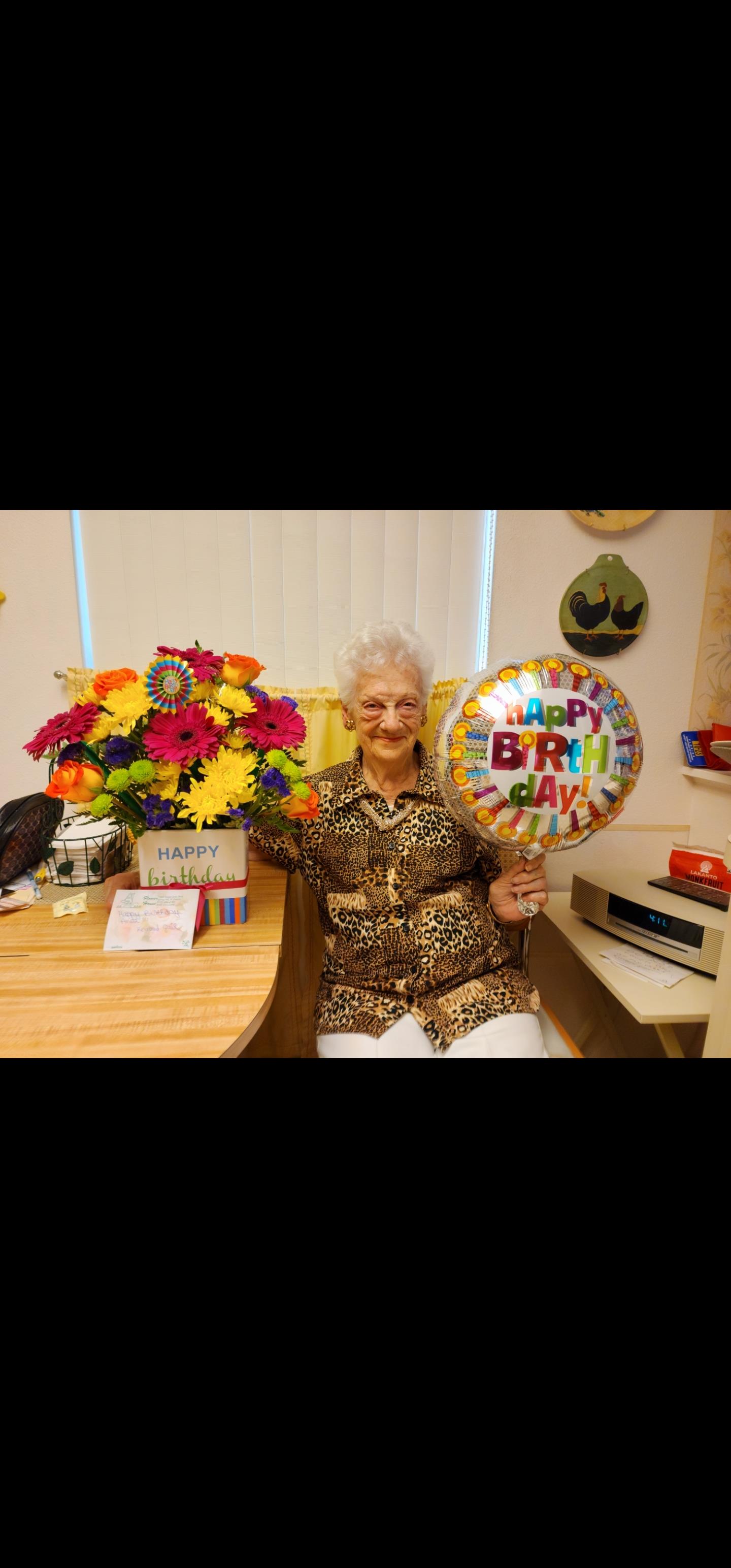 An elderly woman enjoys a birthday celebration holding a balloon and surrounded by colorful flowers.