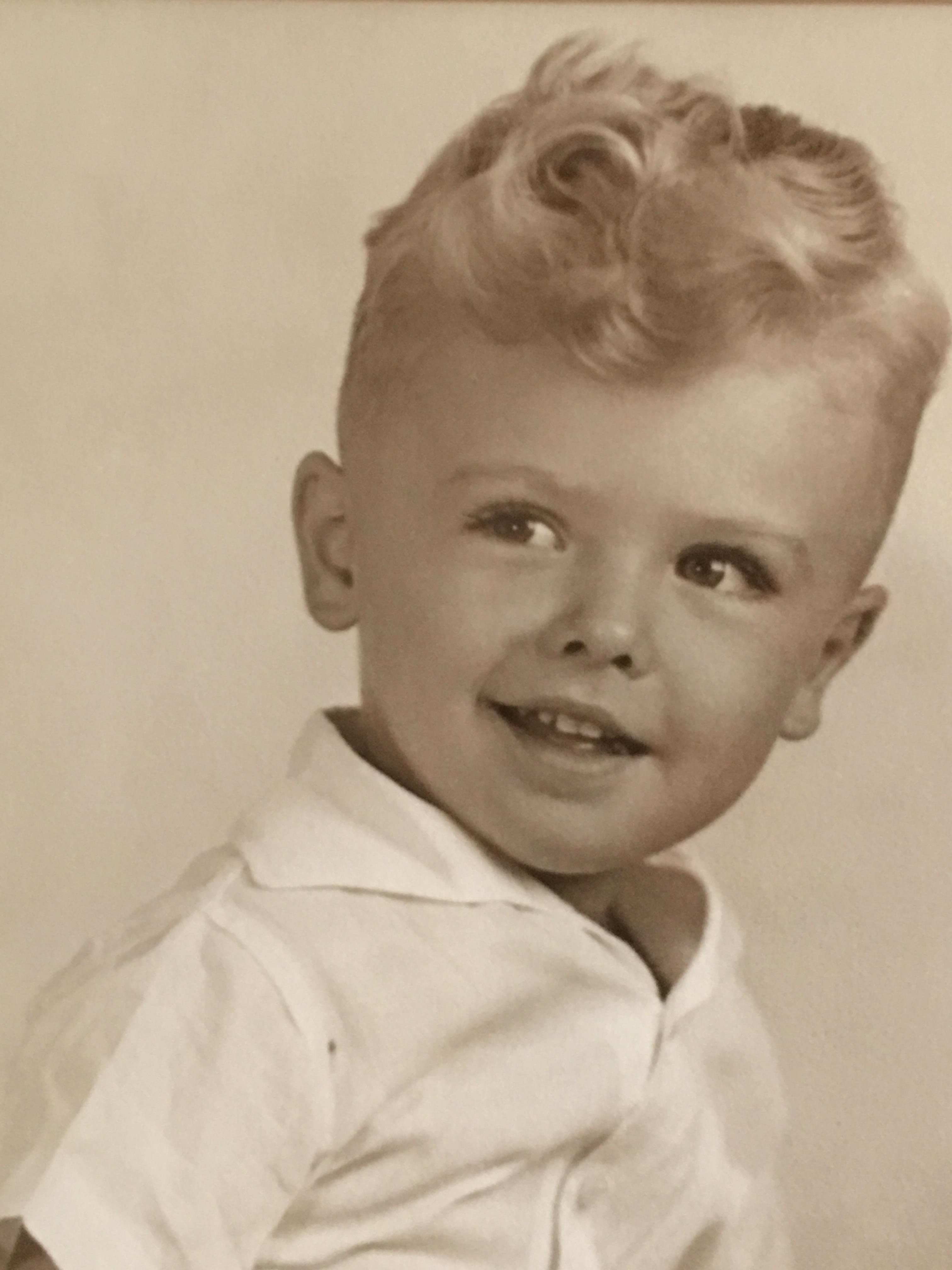 A cheerful young boy with curly blonde hair is smiling playfully, showcasing a vintage charm.