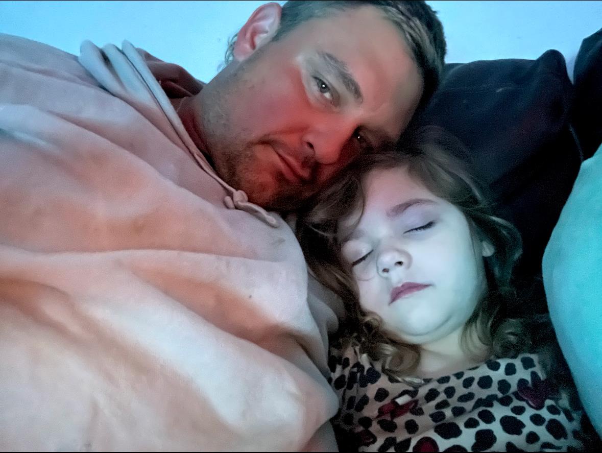 A father enjoys a quiet moment on the couch, holding his daughter who peacefully sleeps beside him.
