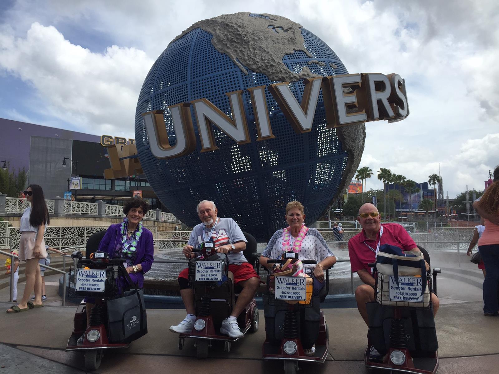 Four friends ride scooters in front of a large globe, capturing a joyful day at the theme park.