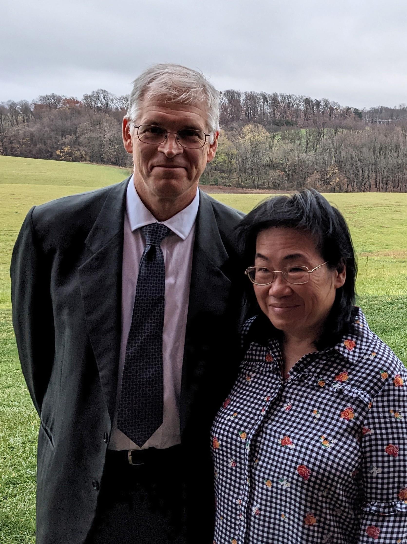 A couple stands together, smiling, against a lush green background during a spring event in nature.