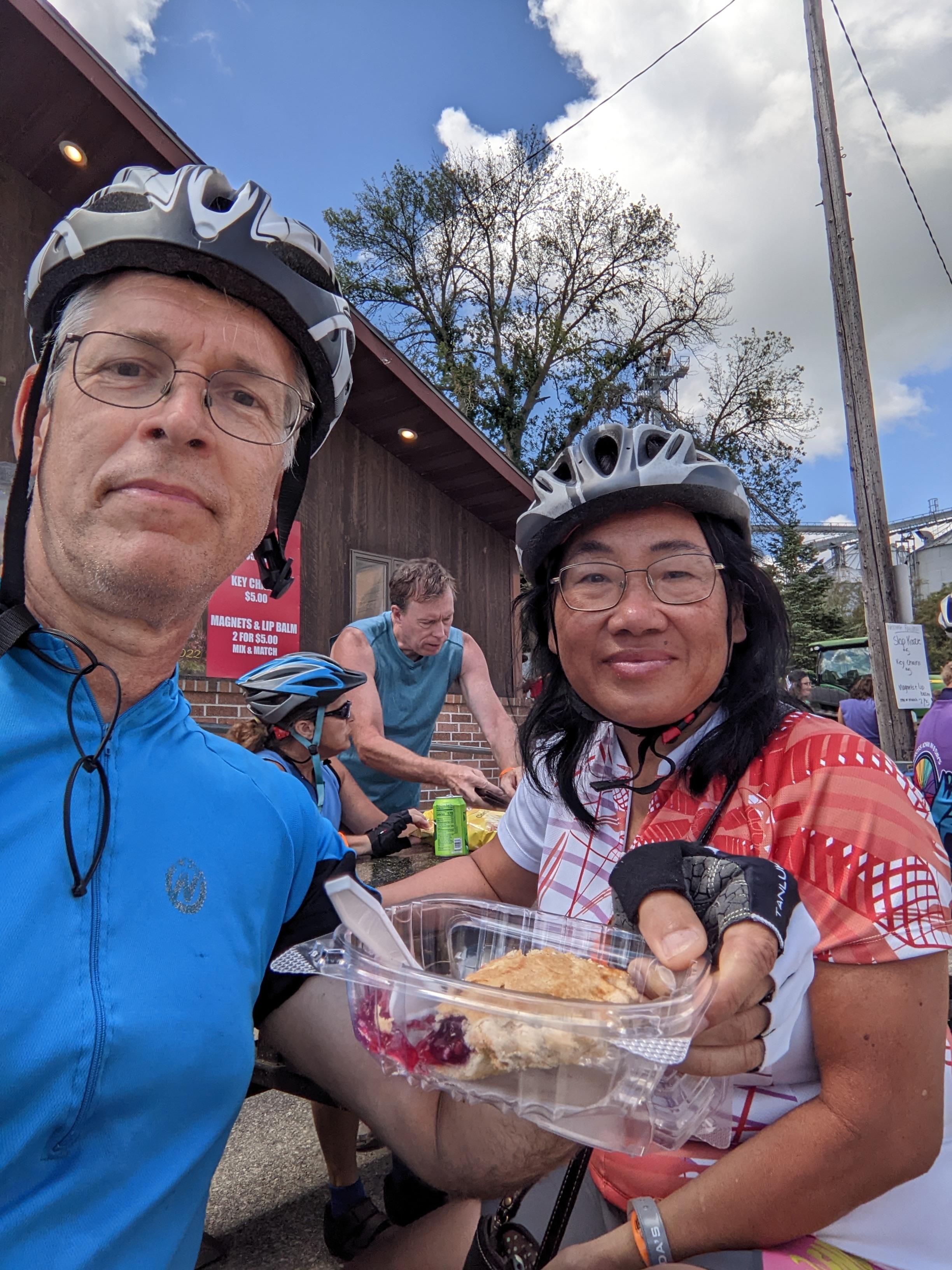 Two cyclists pause for a snack and chat while other riders gather nearby at a scenic spot.