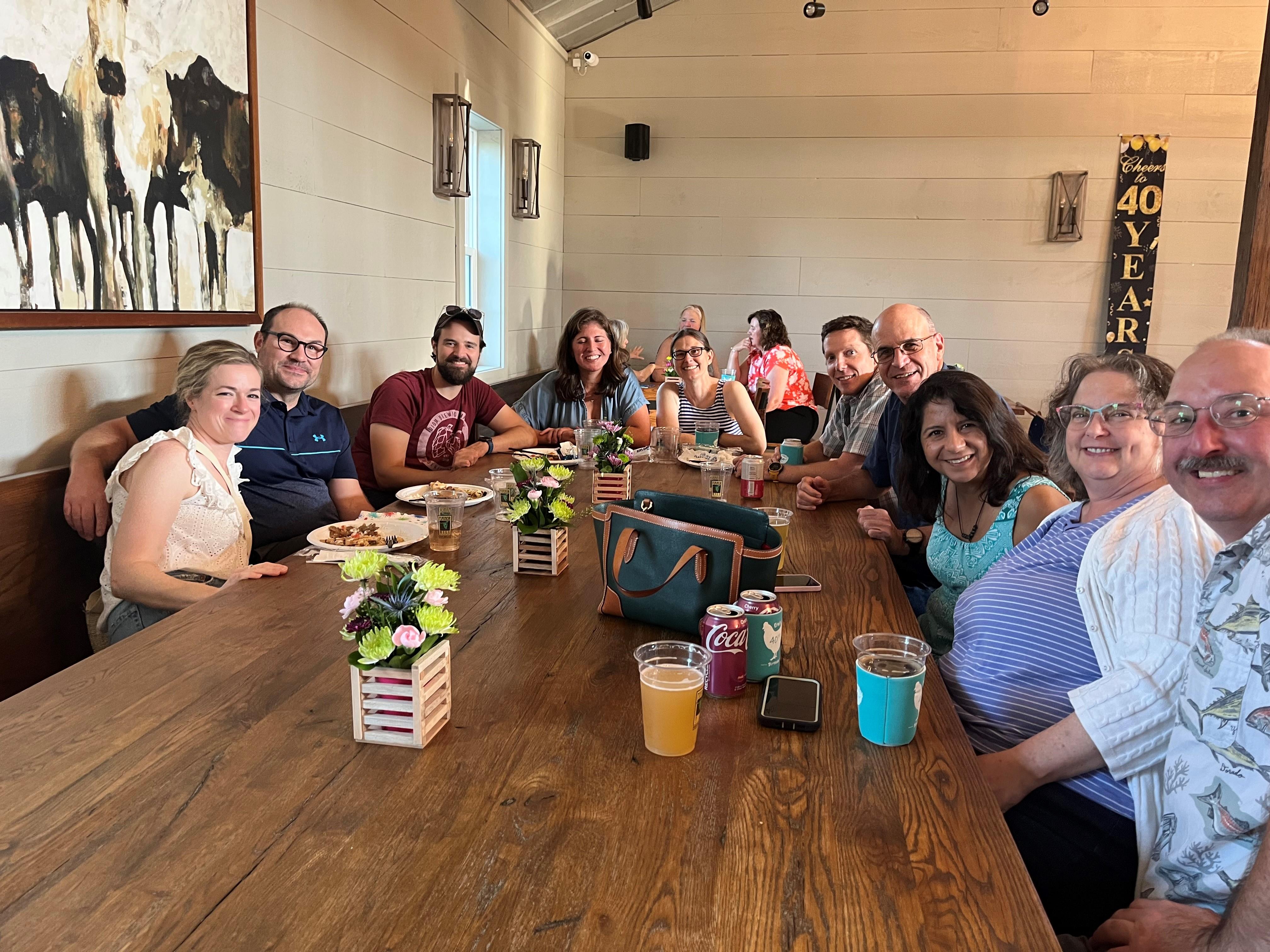 Friends gather around a long wooden table sharing food and drinks in a warm, inviting space.