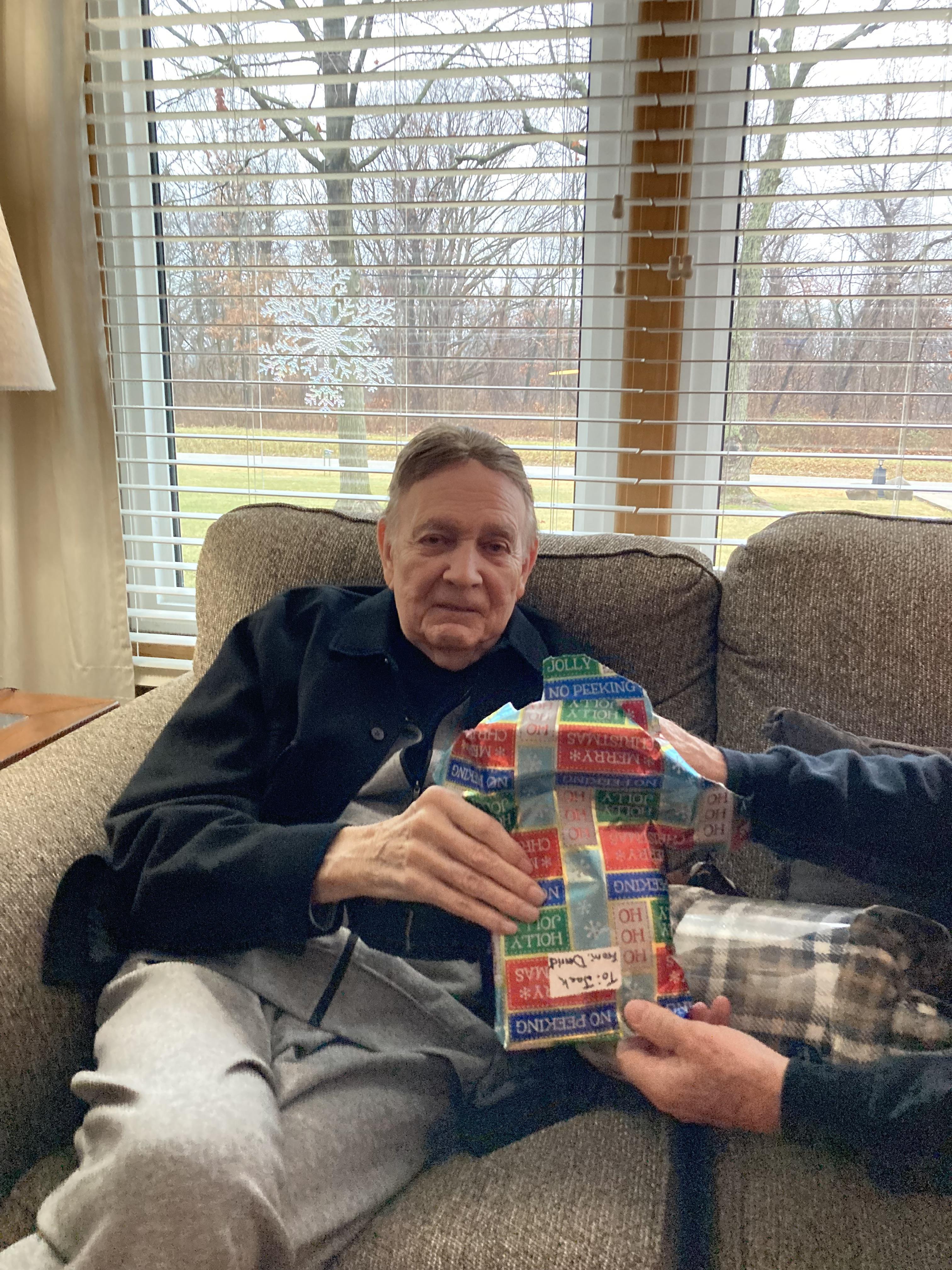 An elderly man enjoys unwrapping a colorful present while seated in a comfortable living space.