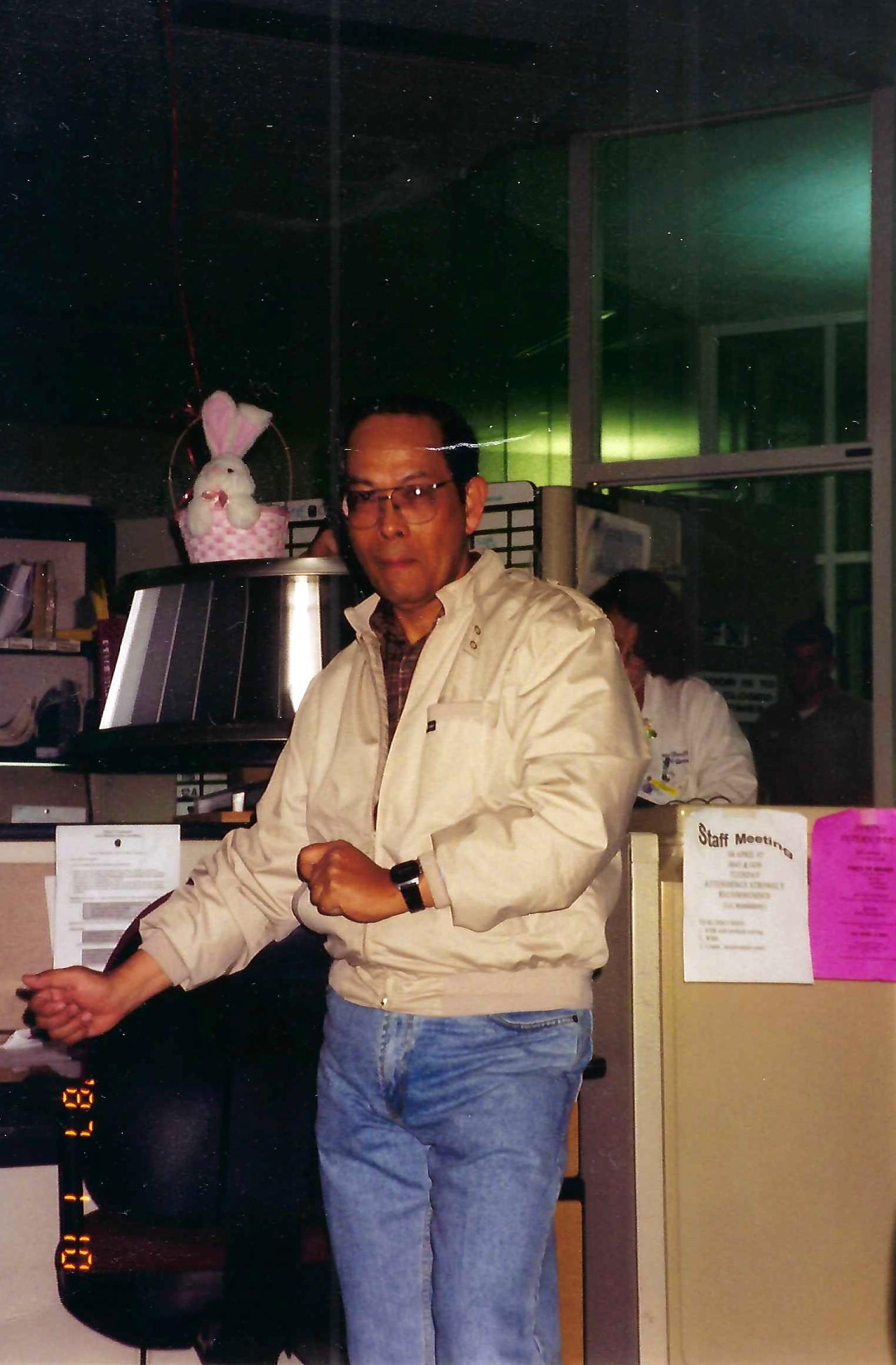 A man wearing glasses and a light jacket poses playfully at his workplace during a lively gathering.