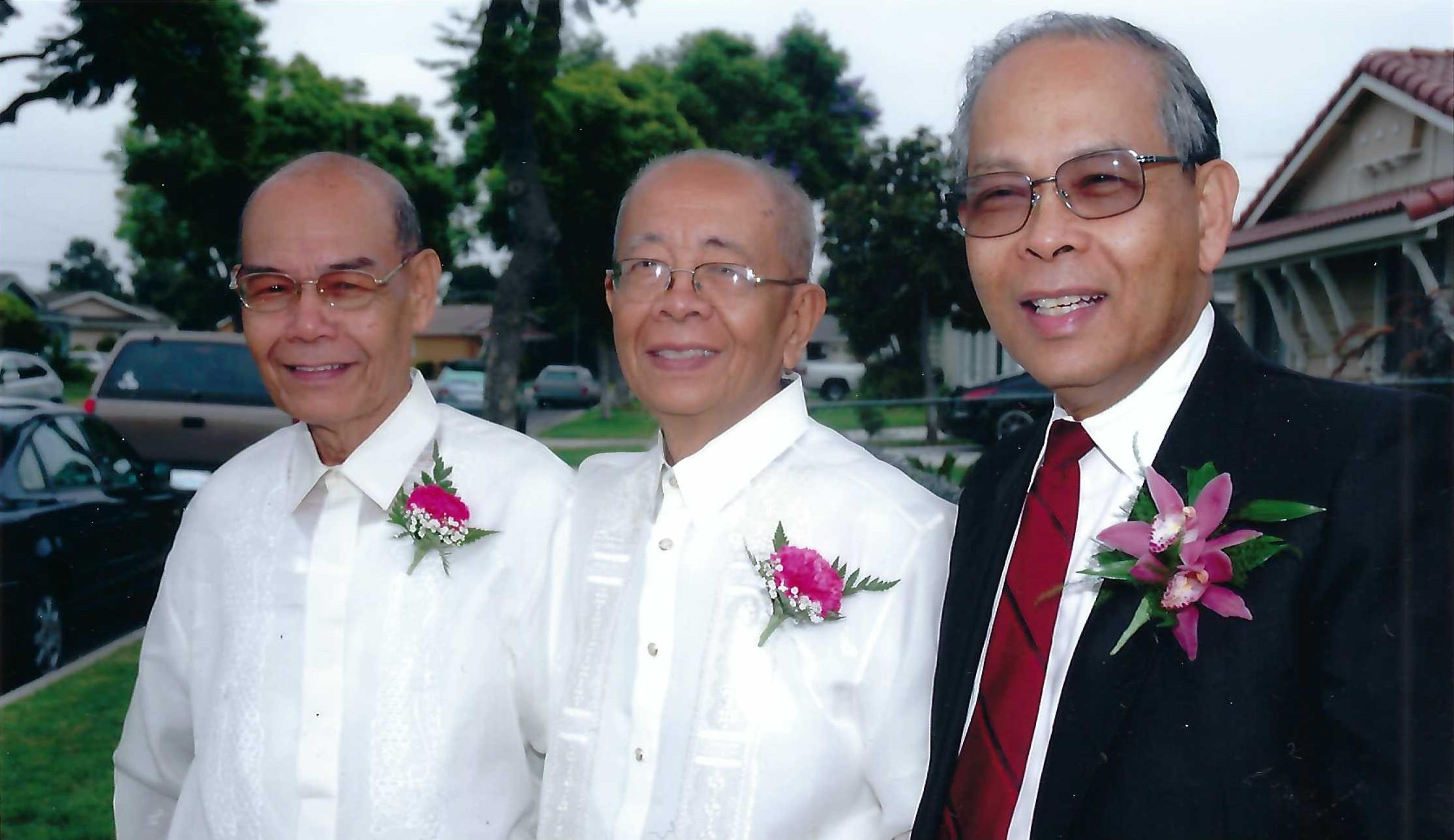 Three older gentlemen wearing traditional attire stand together, smiling in a garden setting.