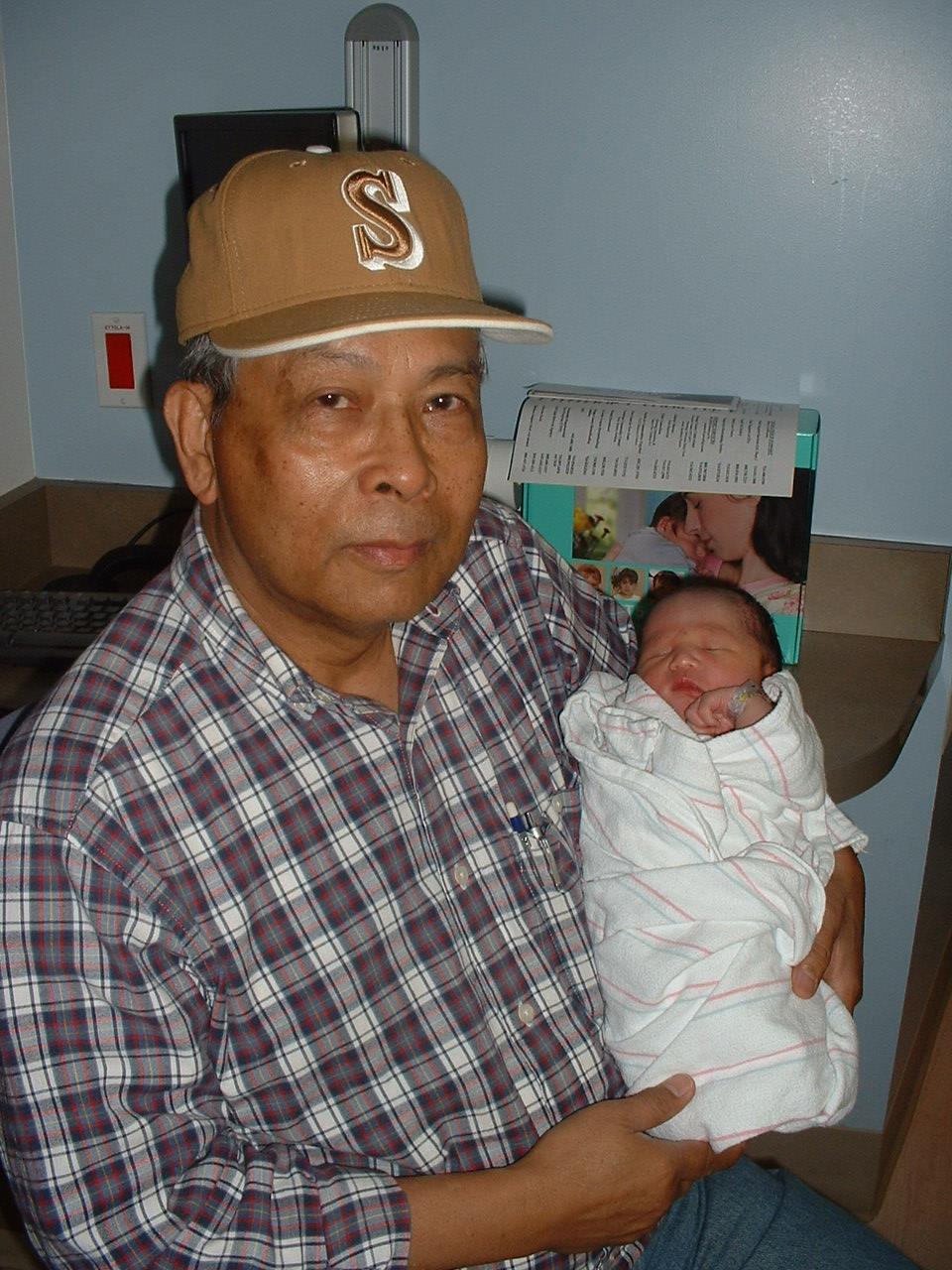 Elderly man smiles while cradling a wrapped newborn baby in a hospital setting.