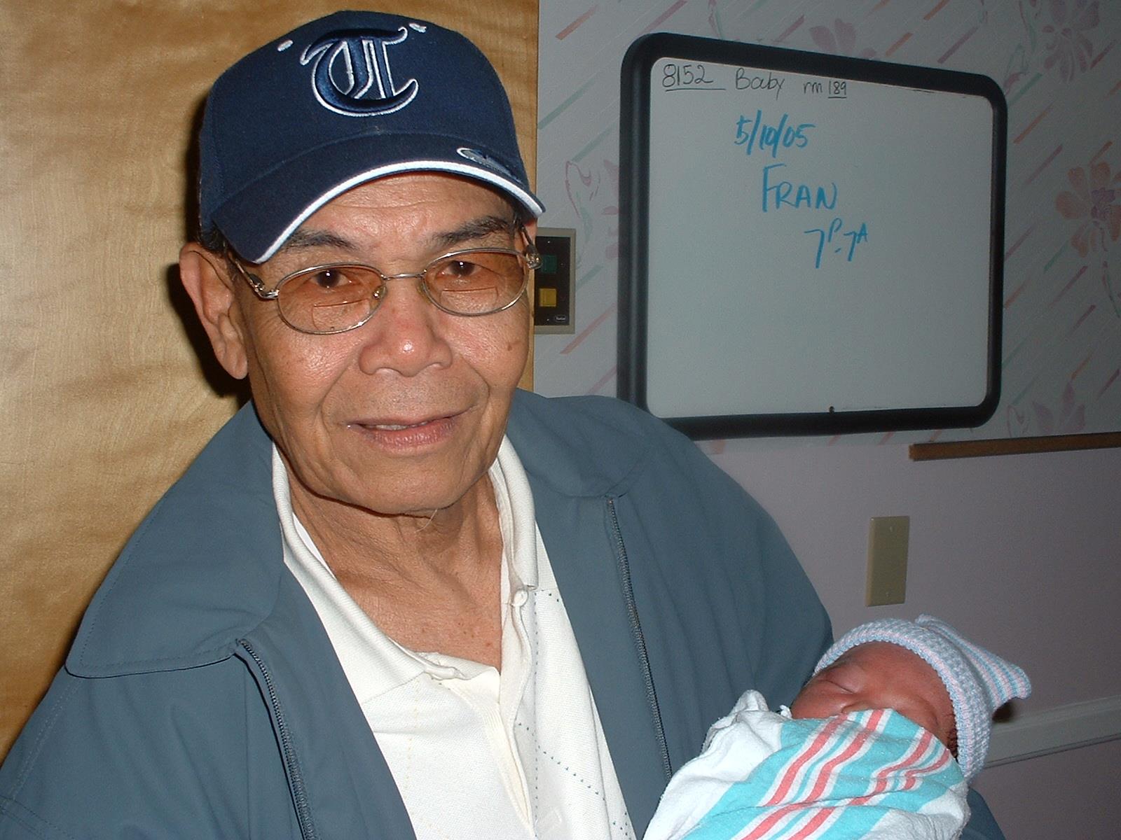 Elderly man holds a baby wrapped in a blanket while smiling in a warm hospital setting.