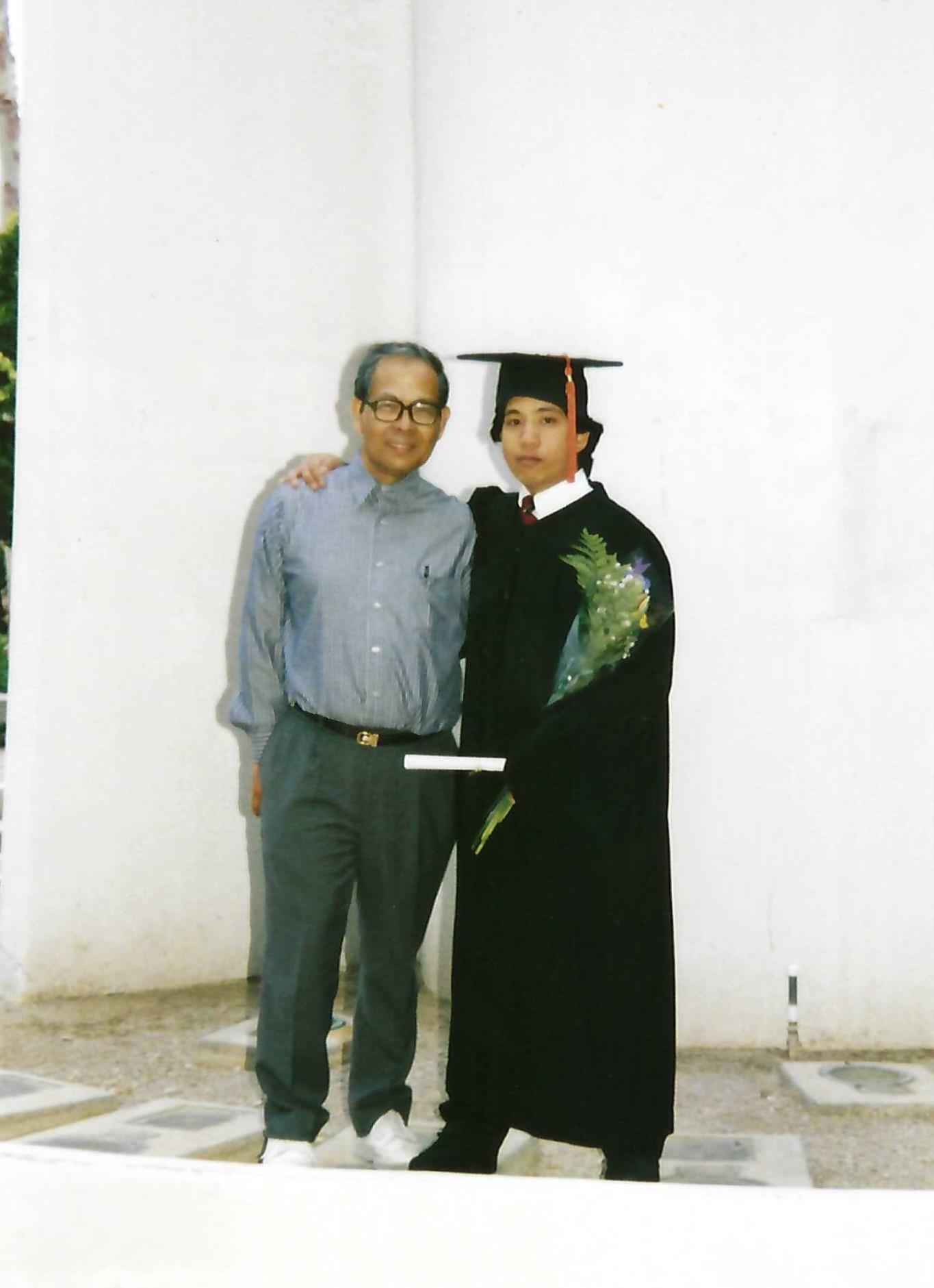 Son in graduation gown stands proudly with father, holding diploma in a courtyard setting.
