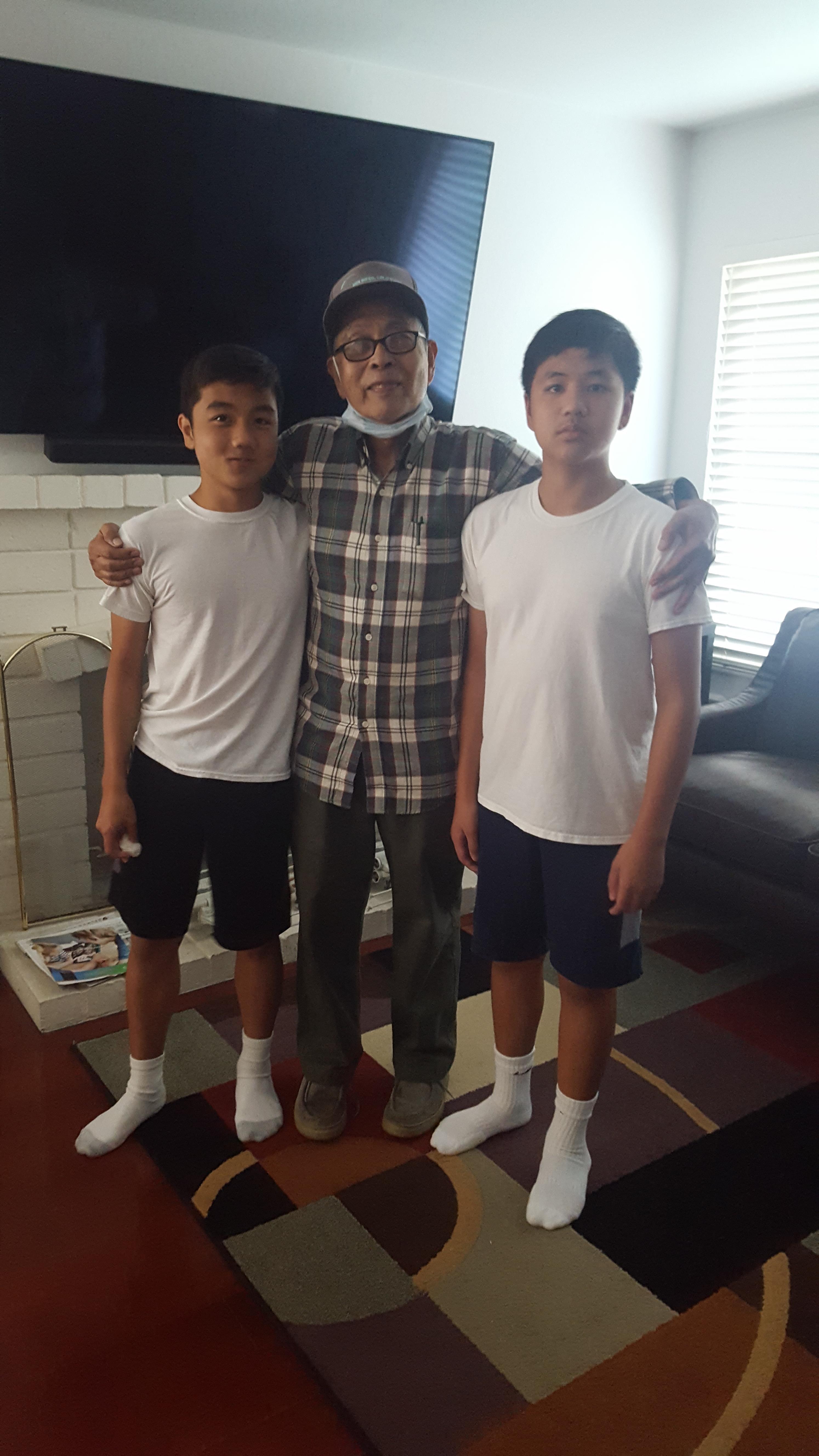 Two boys in white shirts and shorts stand beside an elderly man in glasses, smiling together.