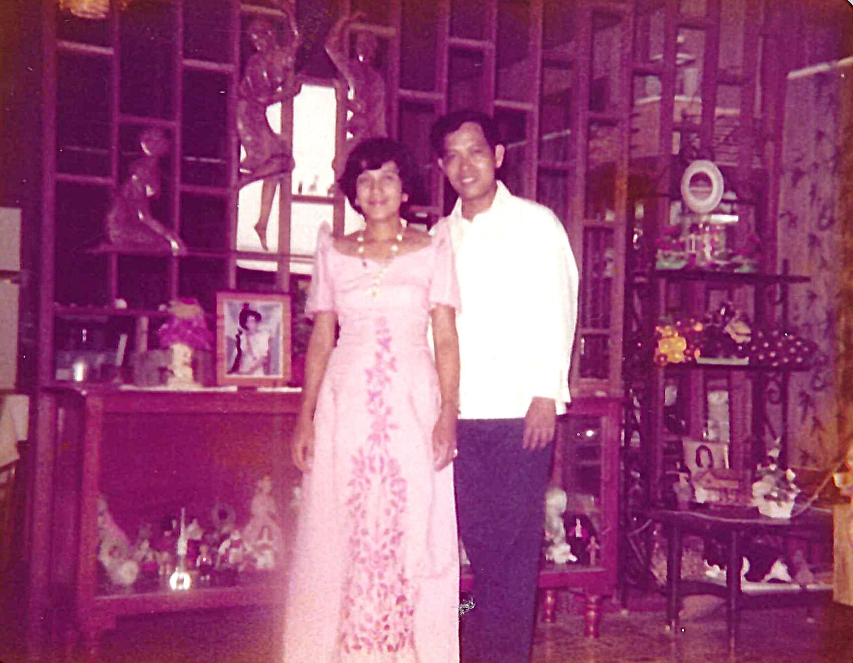A couple stands smiling in traditional clothing, surrounded by various home decorations.