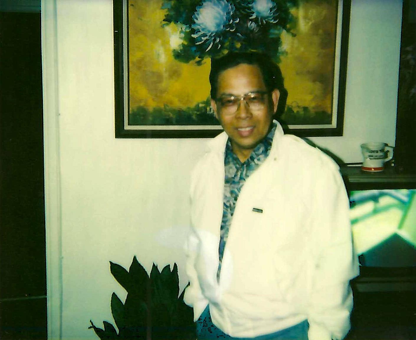 Man with glasses and jacket smiling. Interior setting with plant and artwork.