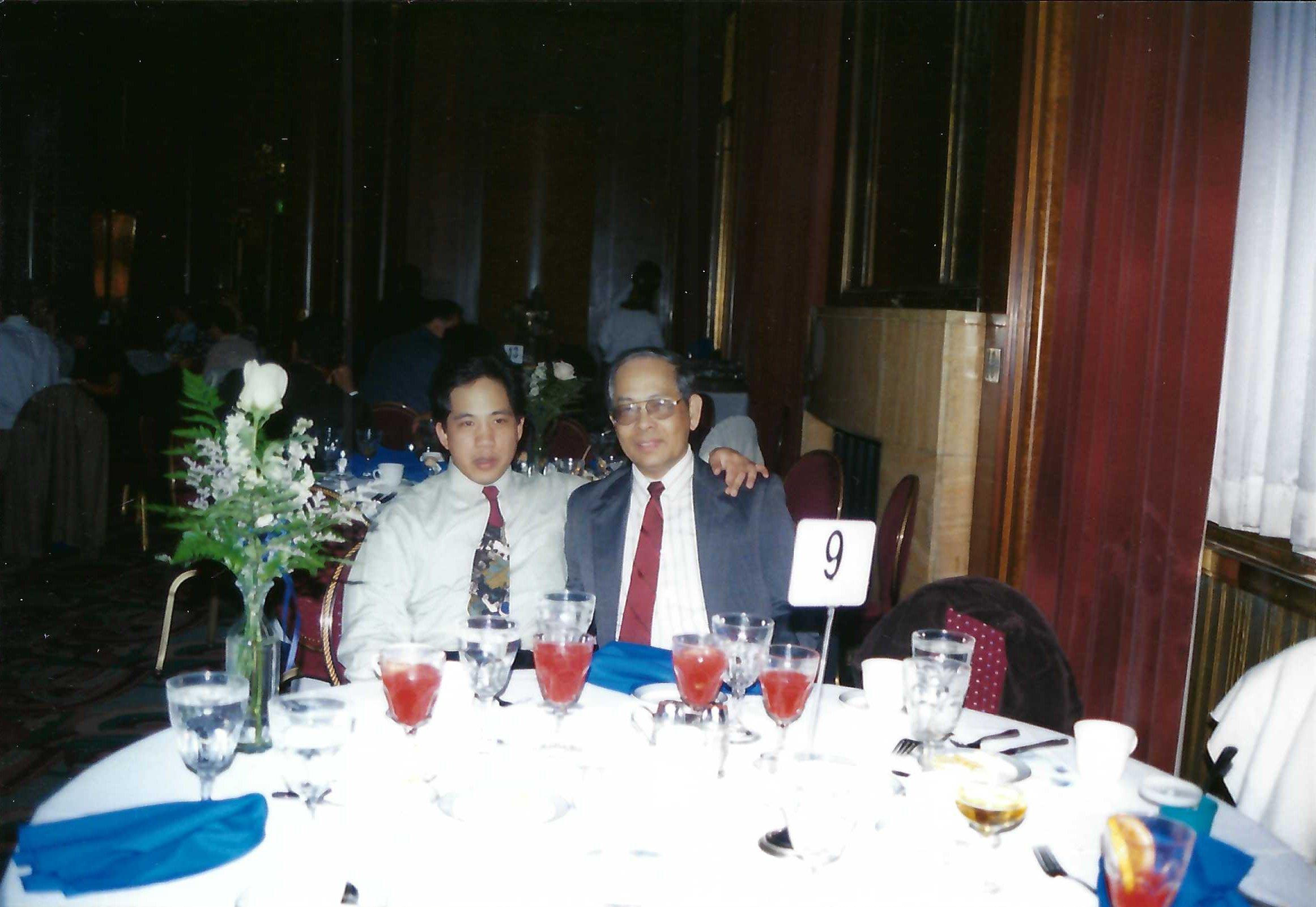 Two individuals seated together at a dinner table adorned with drinks and floral arrangements.