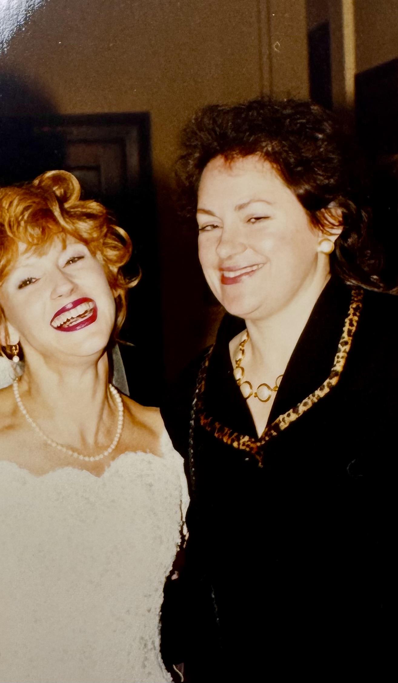 Two women smile warmly while celebrating together at a wedding, showcasing their stylish attire.