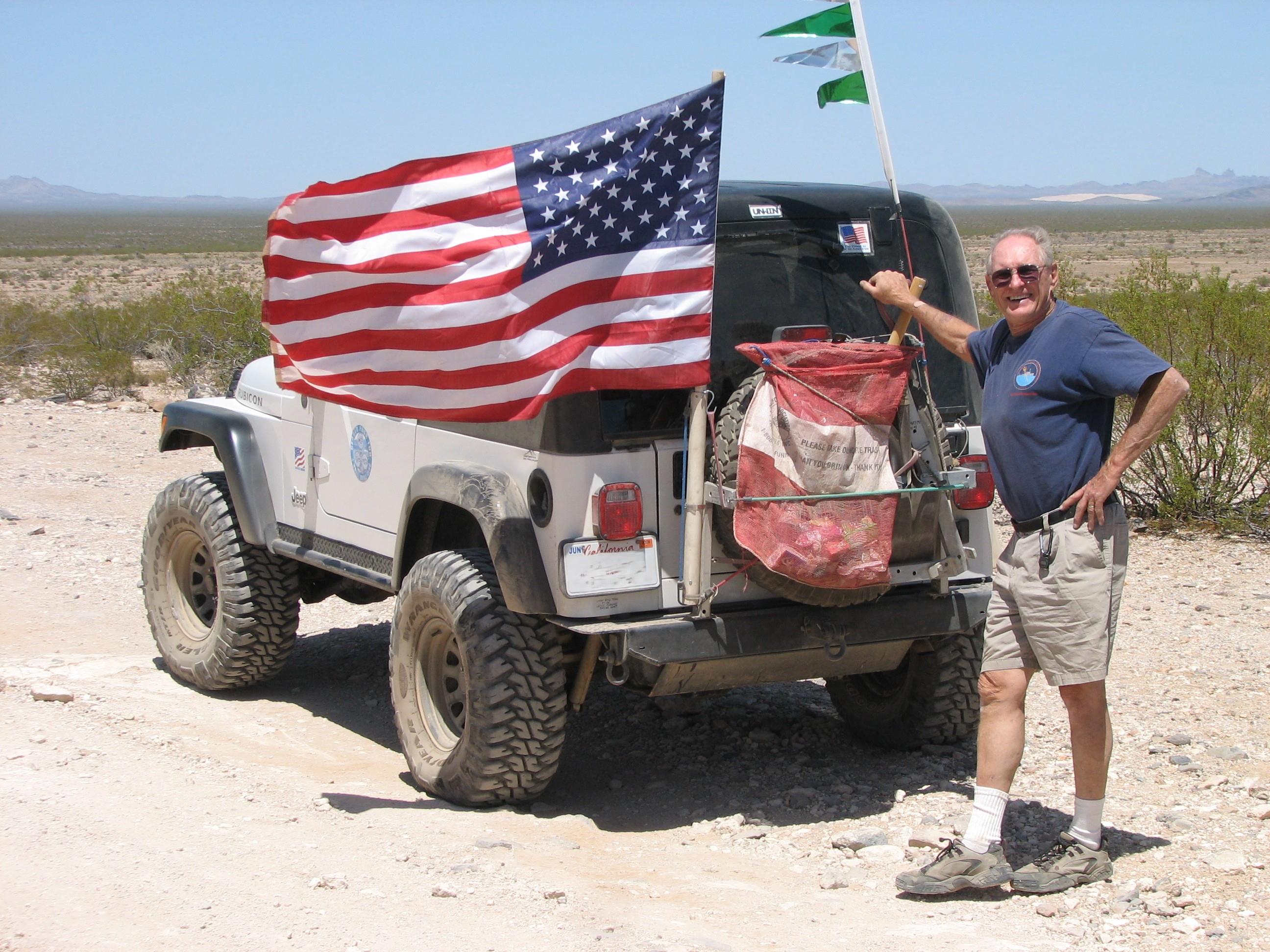 A man stands next to a jeep adorned with an American flag in a vast desert area.