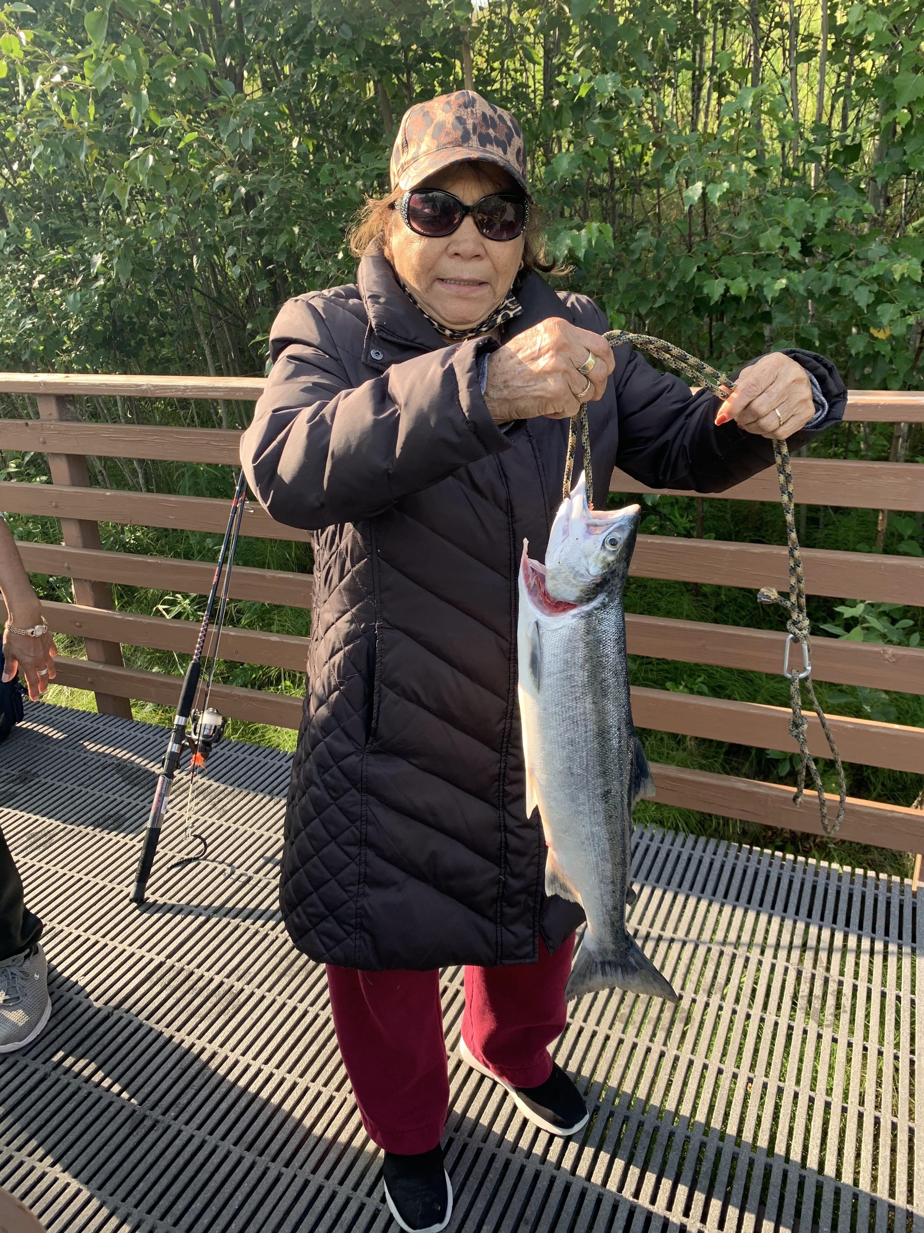 An elderly woman proudly displays a freshly caught salmon while fishing at a riverbank.