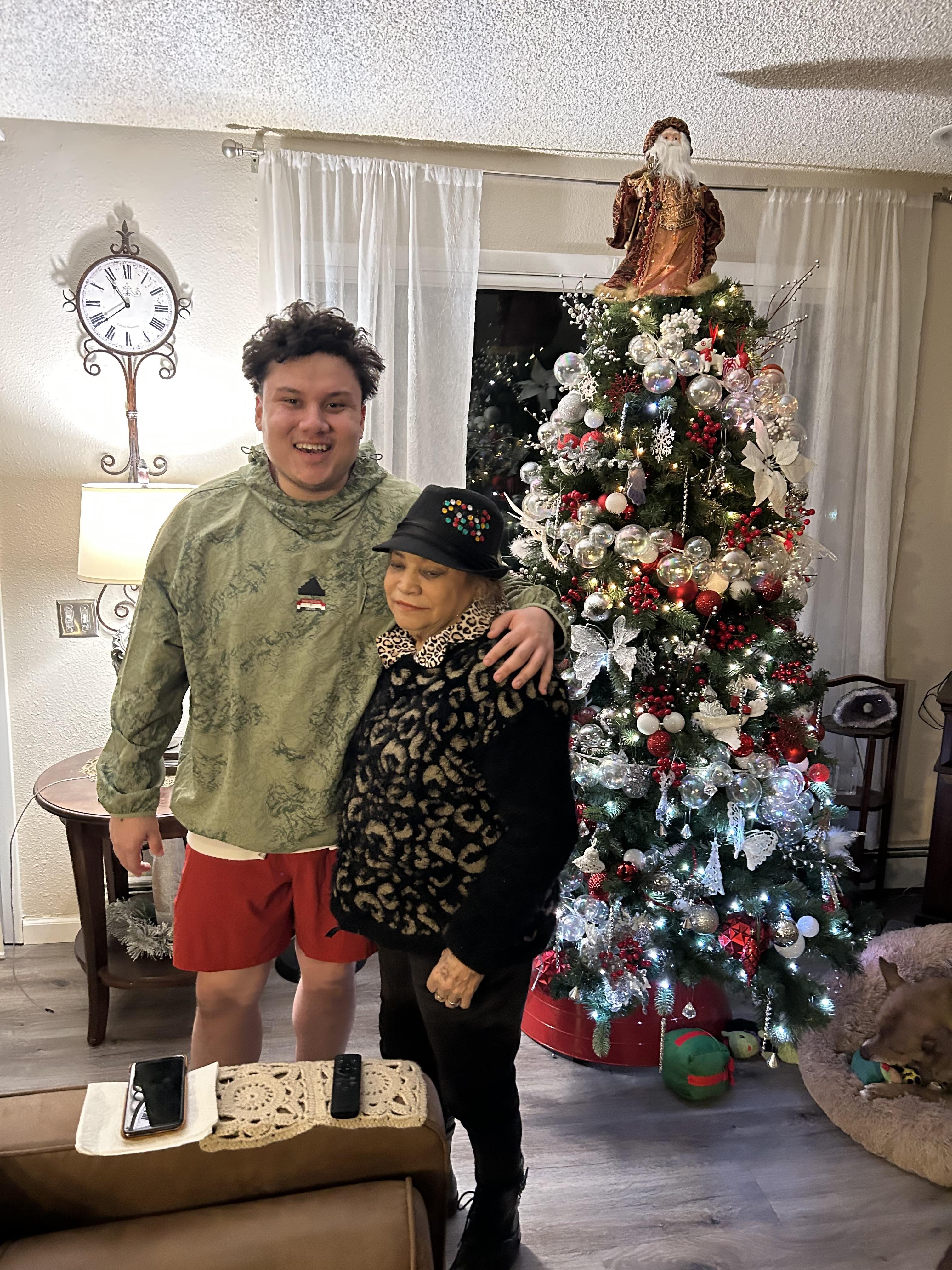 A young man and an elderly woman pose together by a festive Christmas tree in a warm room.