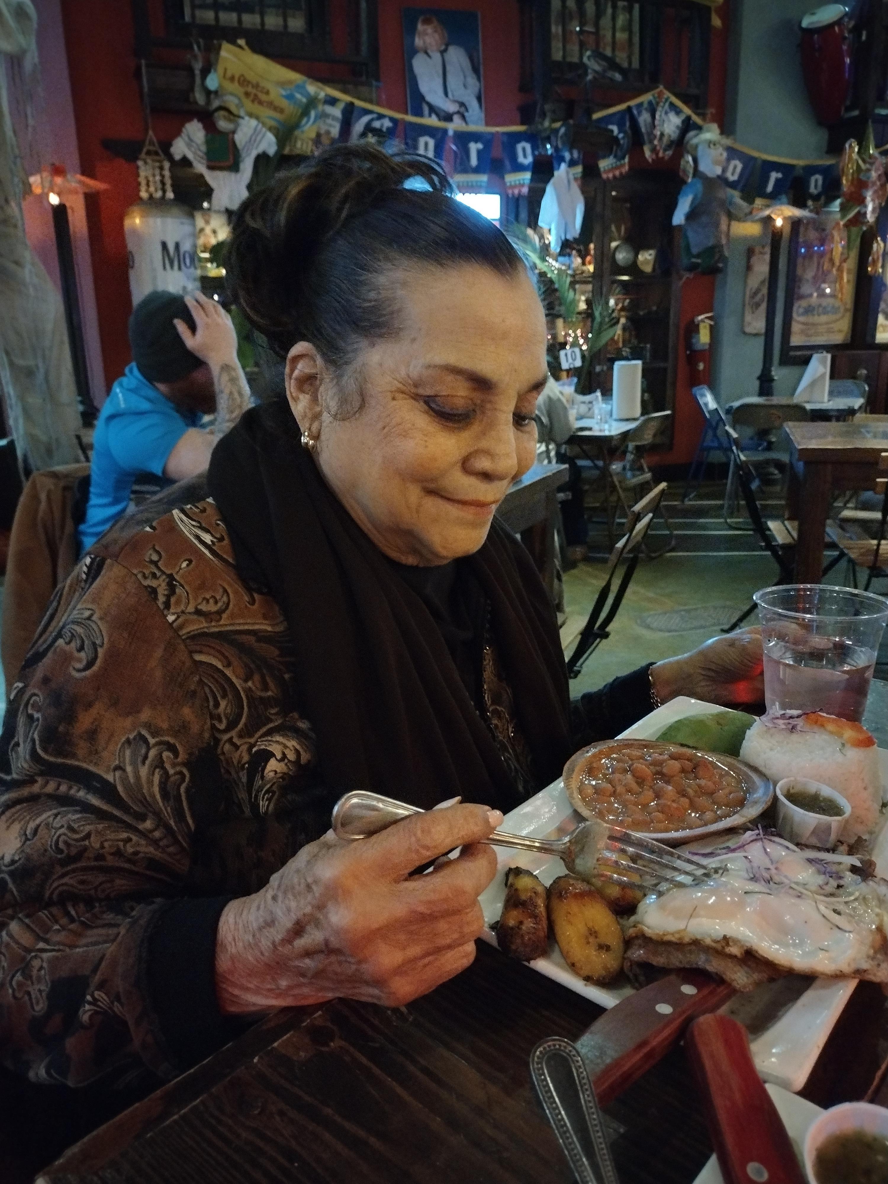 An older woman savors her meal while sitting at a bustling restaurant with warm decorations.