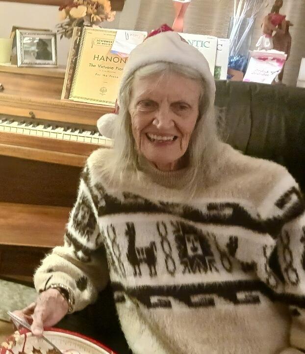 An elderly woman enjoys a cozy winter day, smiling while wearing a festive outfit near a piano.