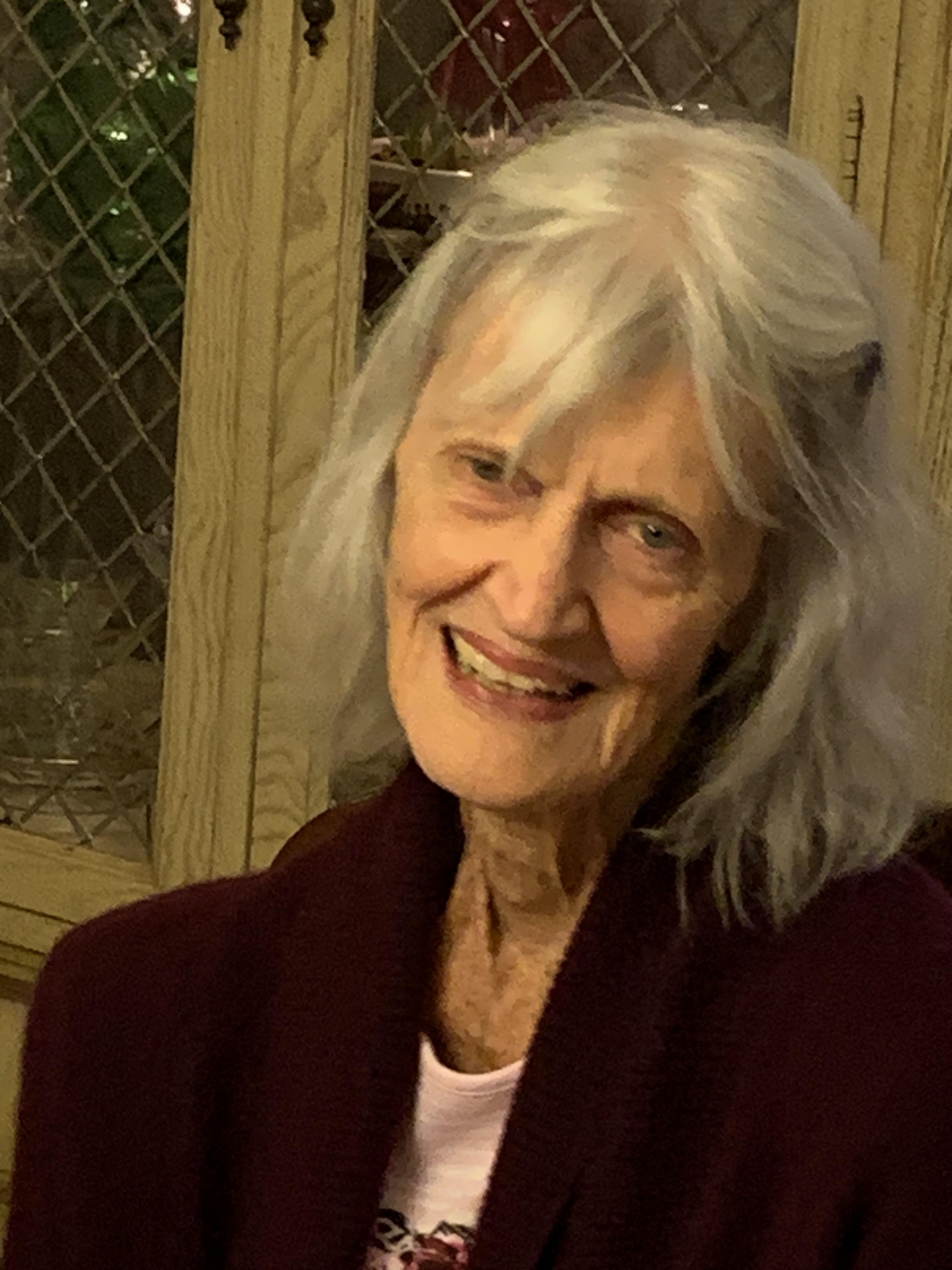 Senior woman with long white hair smiles softly in a warm, inviting indoor environment.