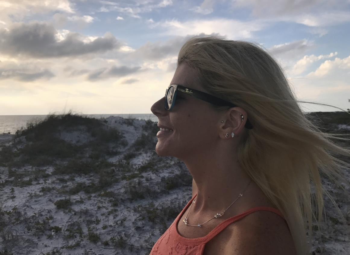 A woman stands on a beach with hair blowing in the wind, enjoying a peaceful moment at sunset.