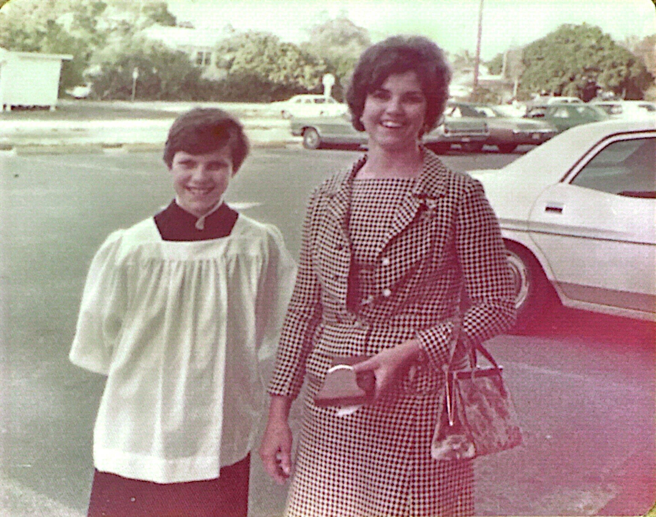 A woman and a young girl smile while dressed in fashionable clothing at a bustling parking lot.