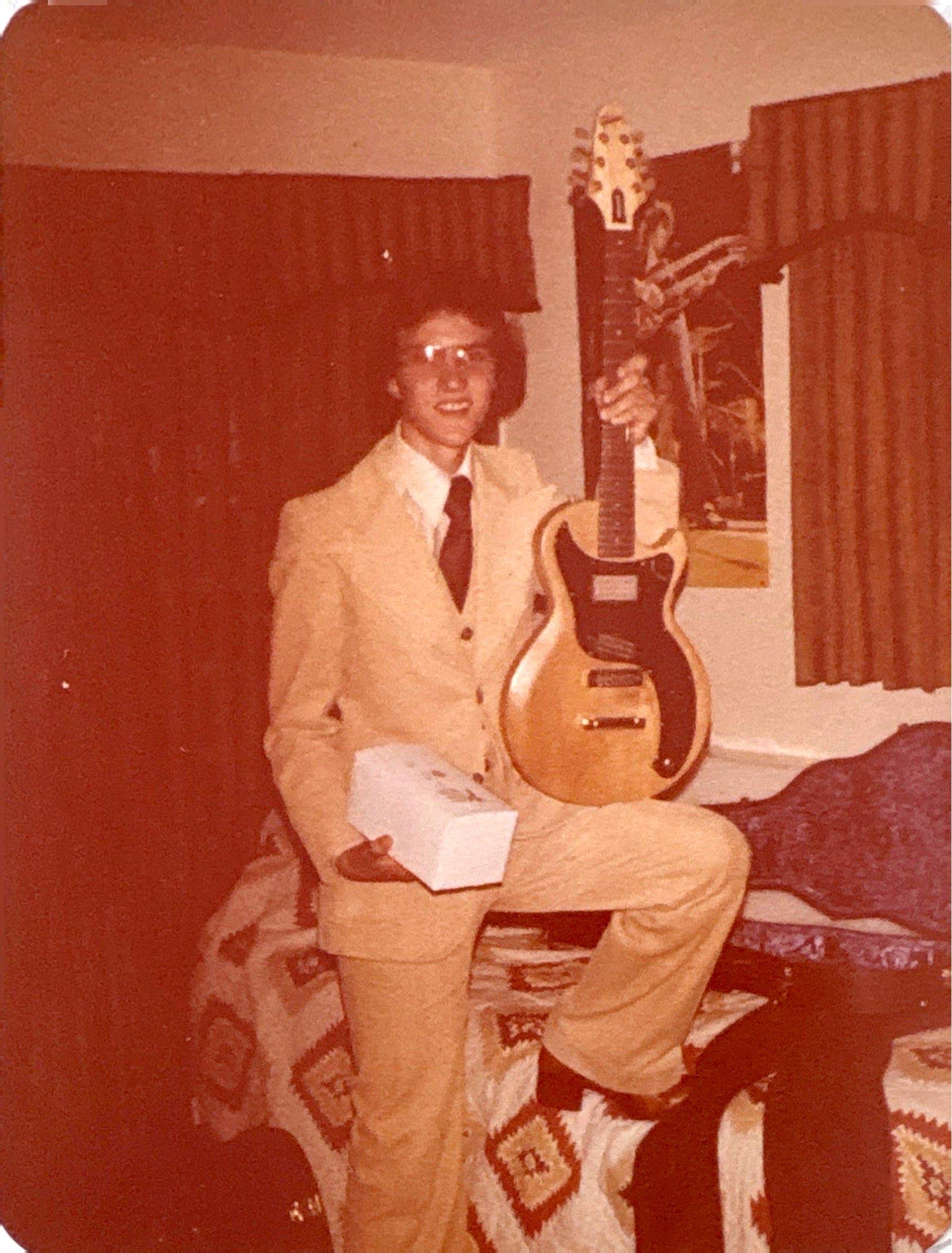 A man in a light suit stands in a cozy room with an electric guitar and a small box.