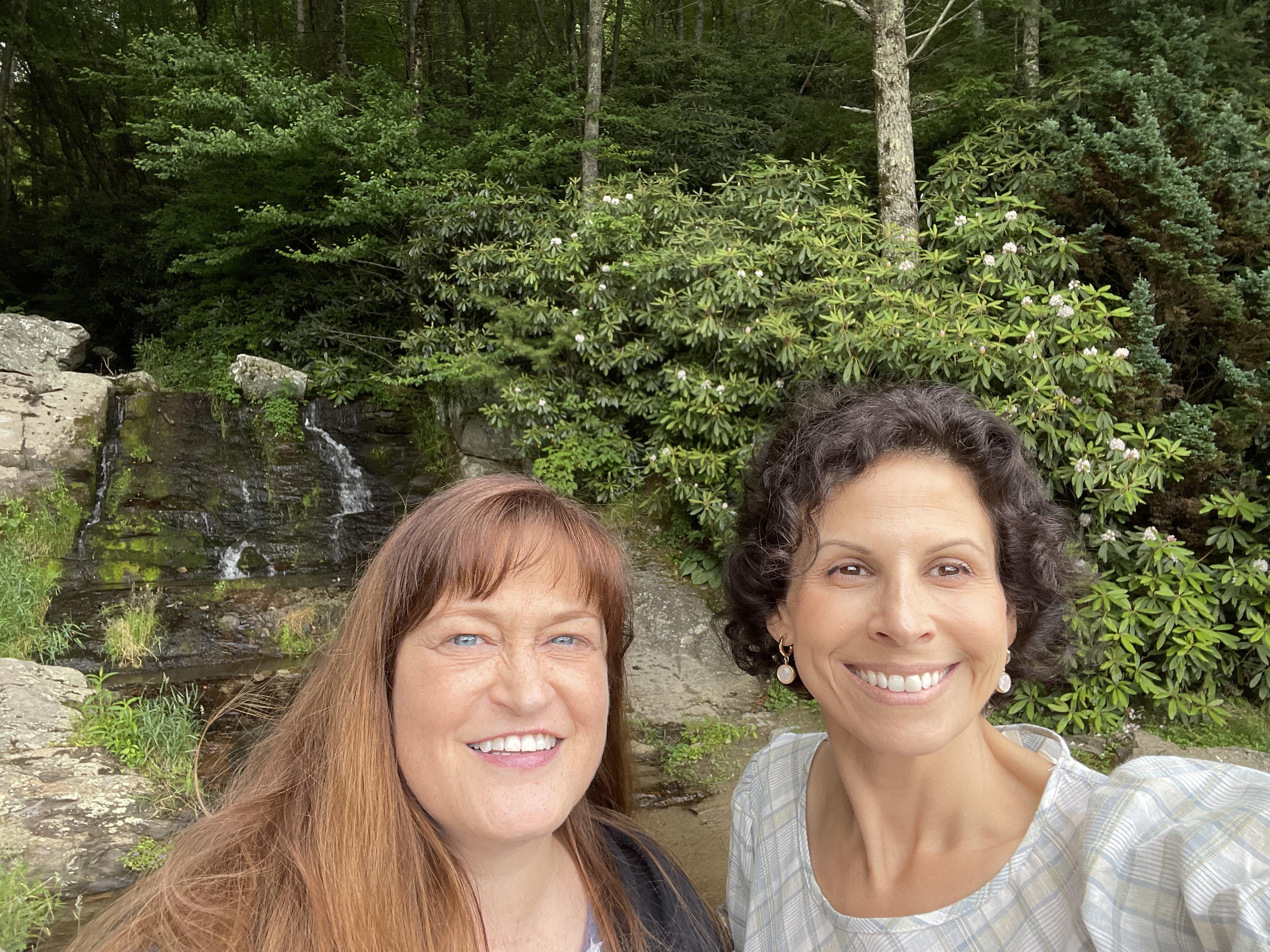 Two friends smile happily as they stand beside a waterfall surrounded by dense greenery.
